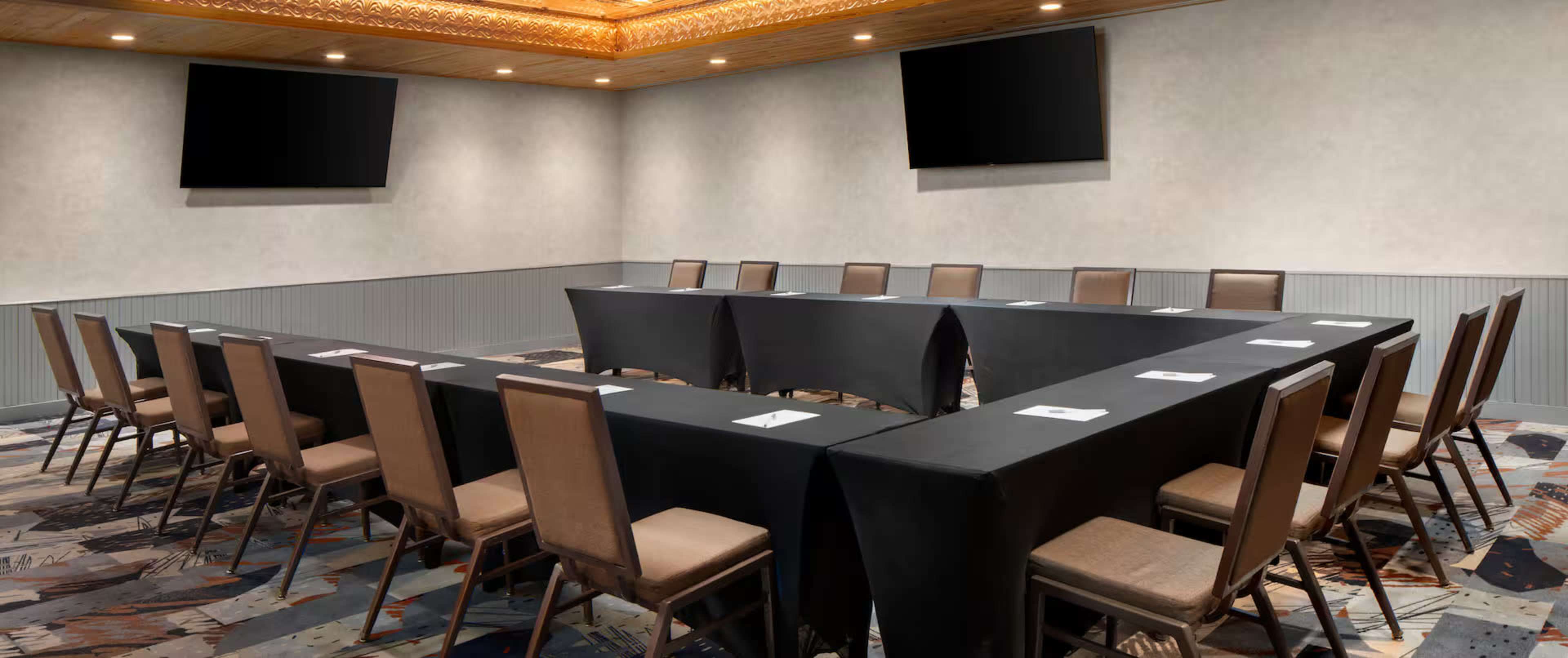 A conference room features a large, U-shaped arrangement of black tables surrounded by chairs, with two screens mounted on the walls.