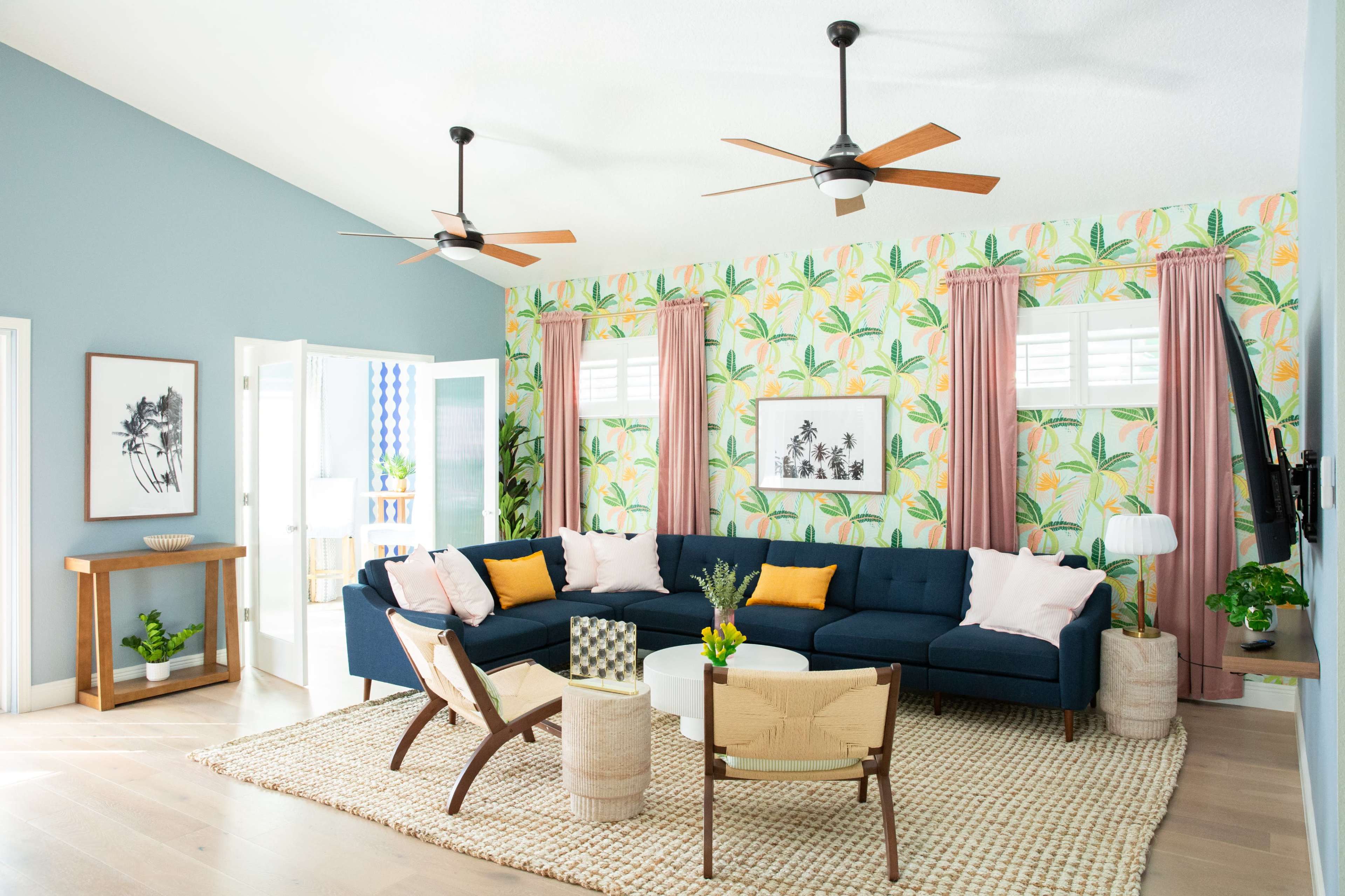 The image shows a bright living room with a blue sectional couch, tropical wallpaper, wooden ceiling fans, and a woven rug.