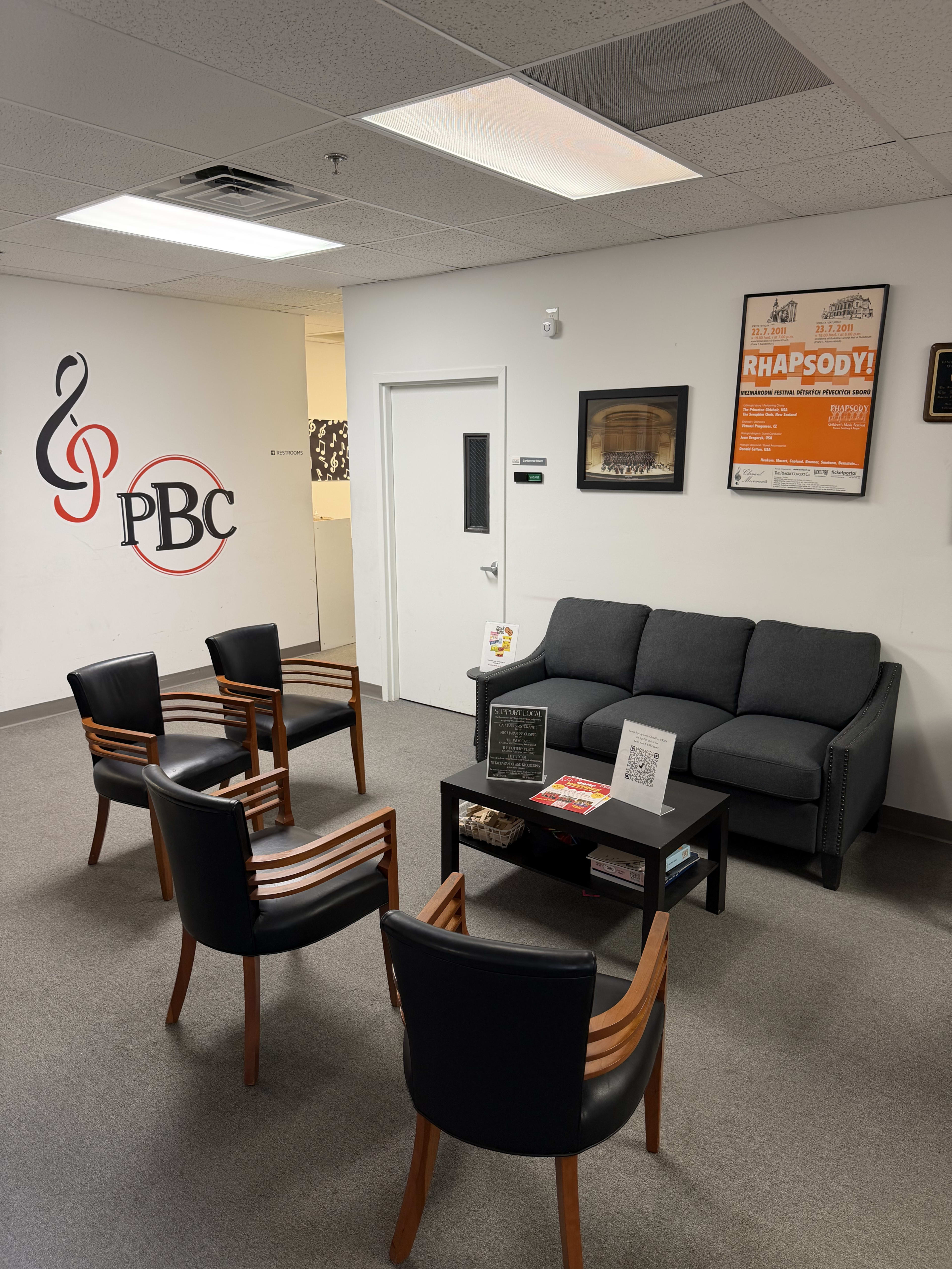 The image shows a waiting area with a gray couch, four black chairs, a coffee table, and various wall decorations including a musical note logo and framed posters.