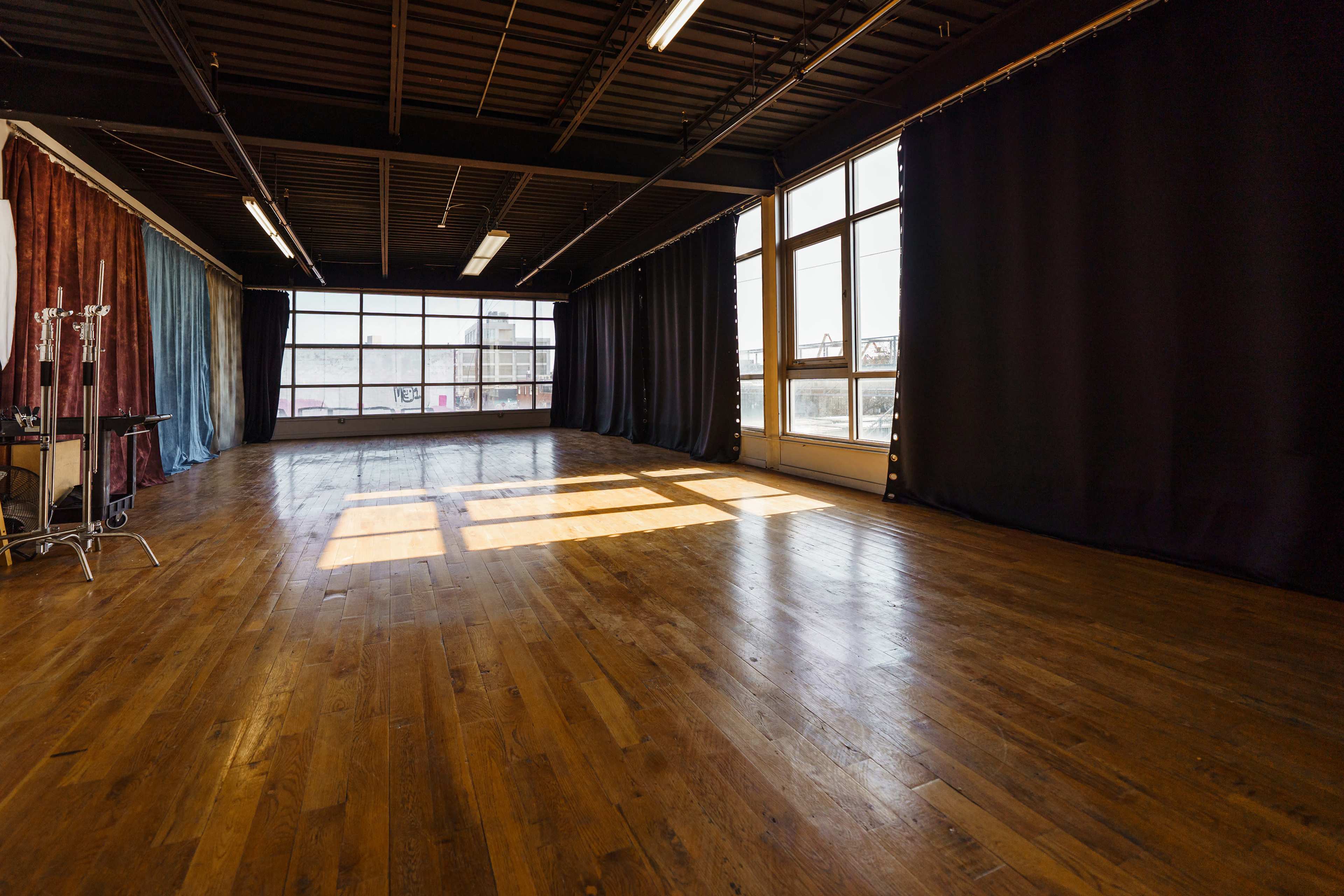 An empty studio space with large windows, wooden floors, and black curtains along the walls.