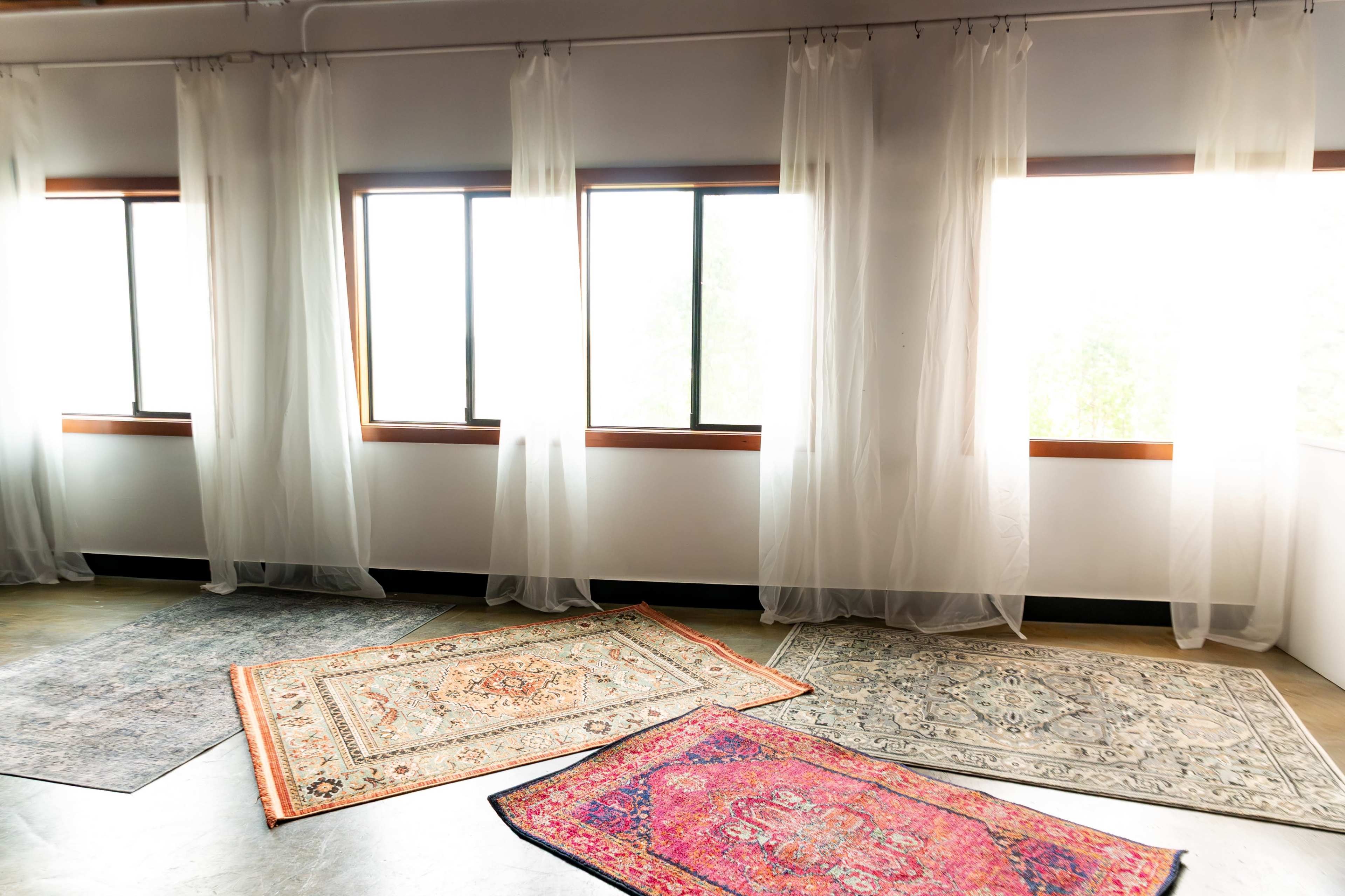 The image shows a bright room with large windows covered by sheer curtains and several decorative rugs spread out on the floor.