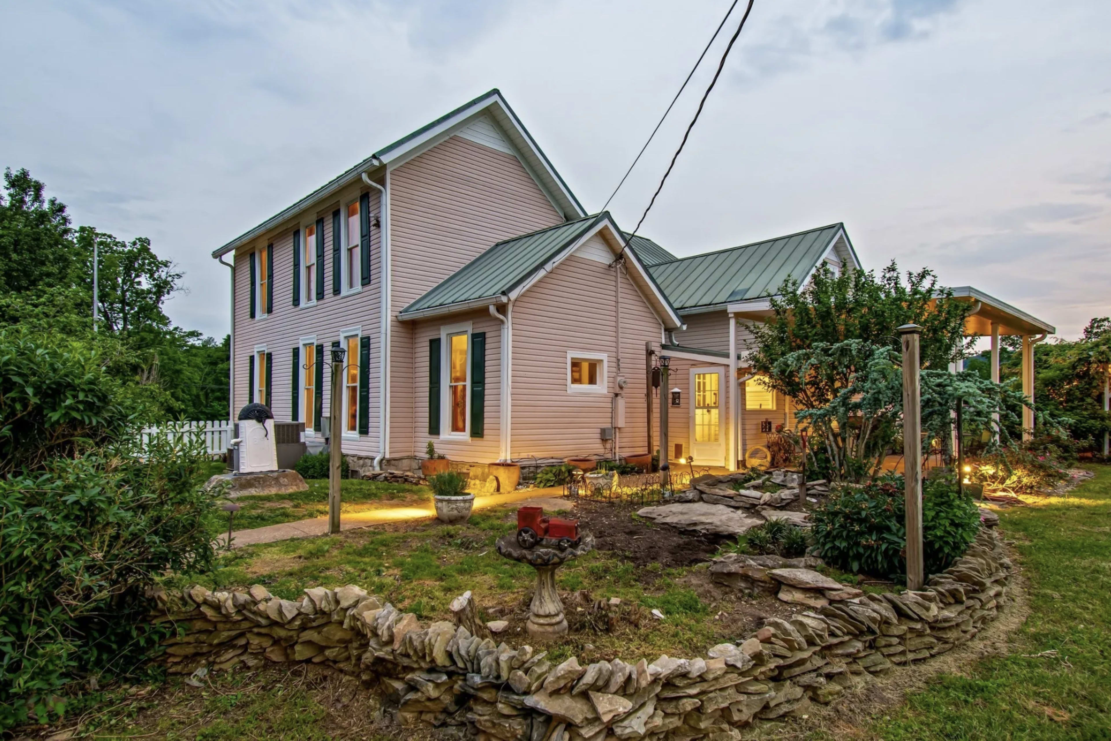The image shows a two-story pink house with a green metal roof, surrounded by landscaped gardens and stone pathways.