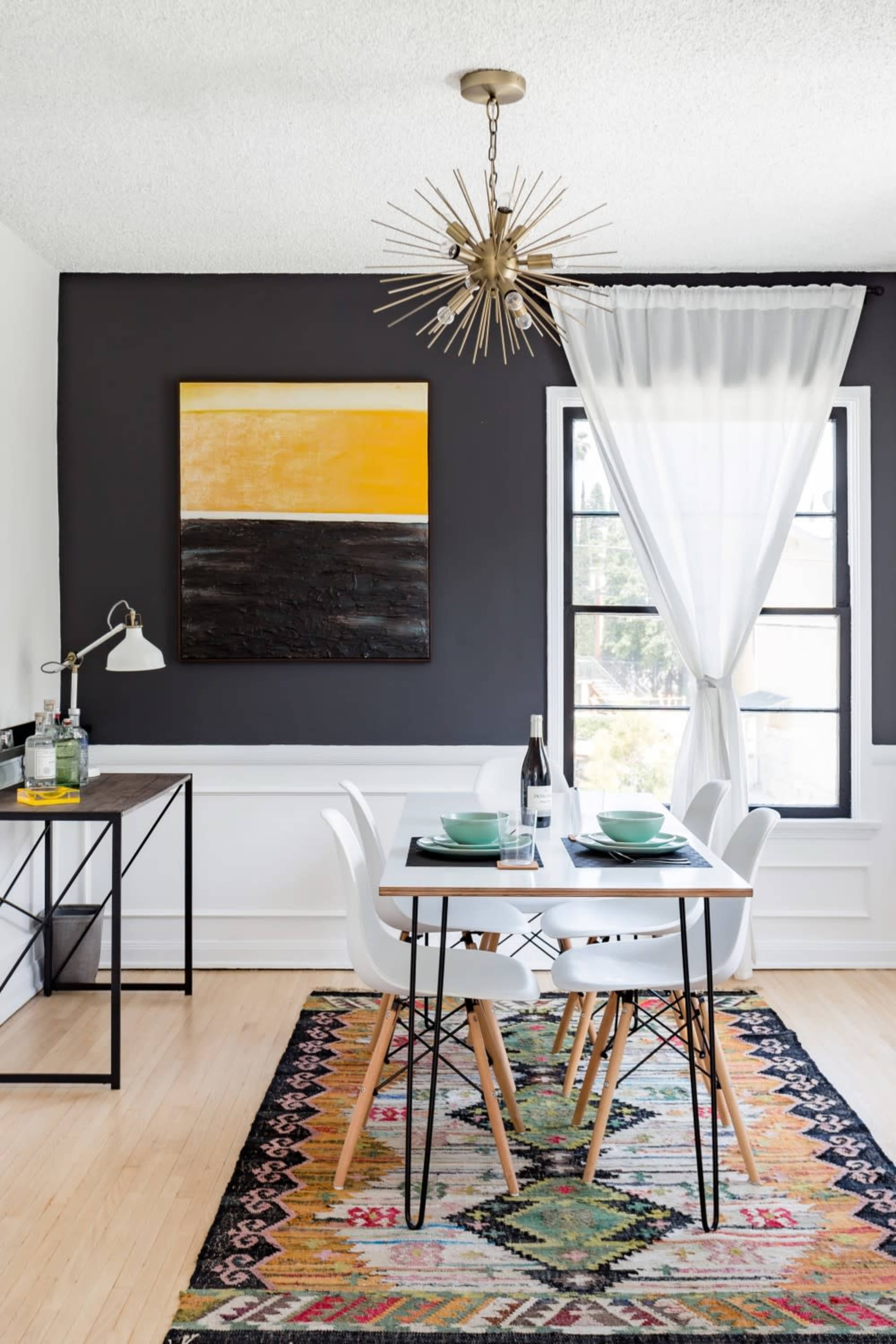 A dining area features a table set for four, accompanied by a patterned rug, a modern light fixture, and a large abstract painting on the wall.