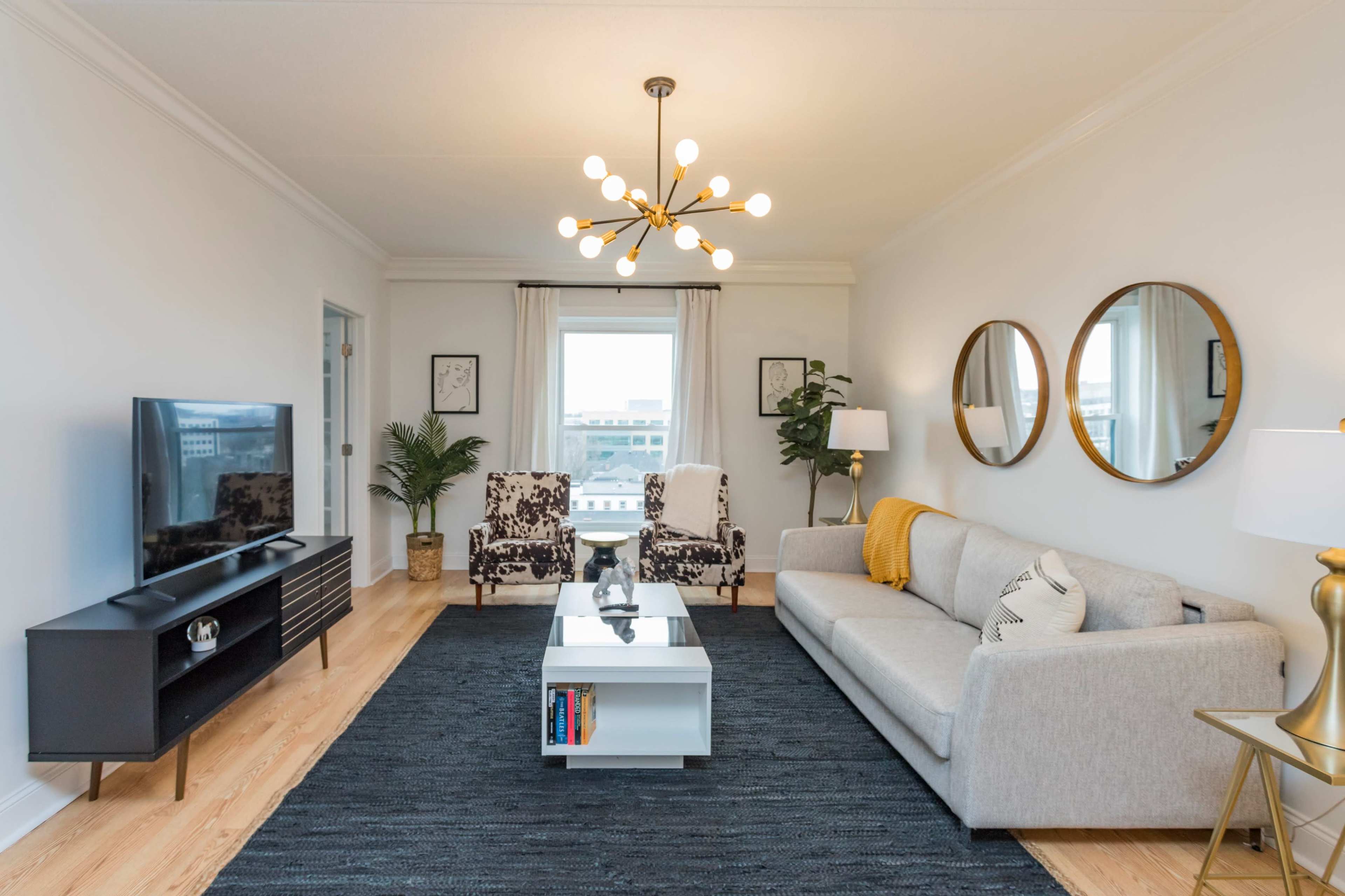 A modern living room features a light gray sofa, two patterned armchairs, a television on a low cabinet, and a coffee table in front of a large window, adorned with decorative mirrors and plants.