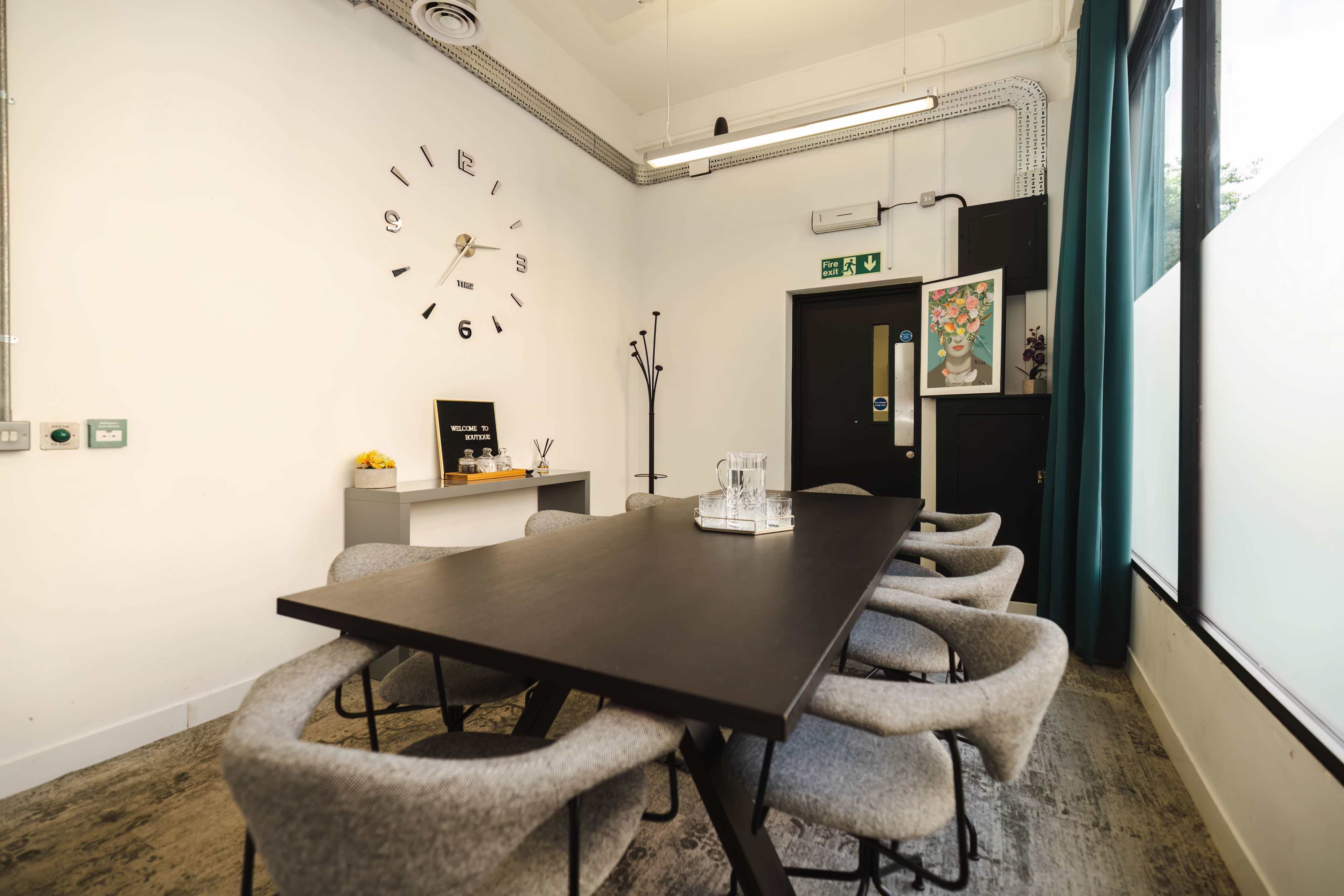 A conference room is equipped with a long black table surrounded by gray chairs, with a wall clock and decorative artwork in the background.