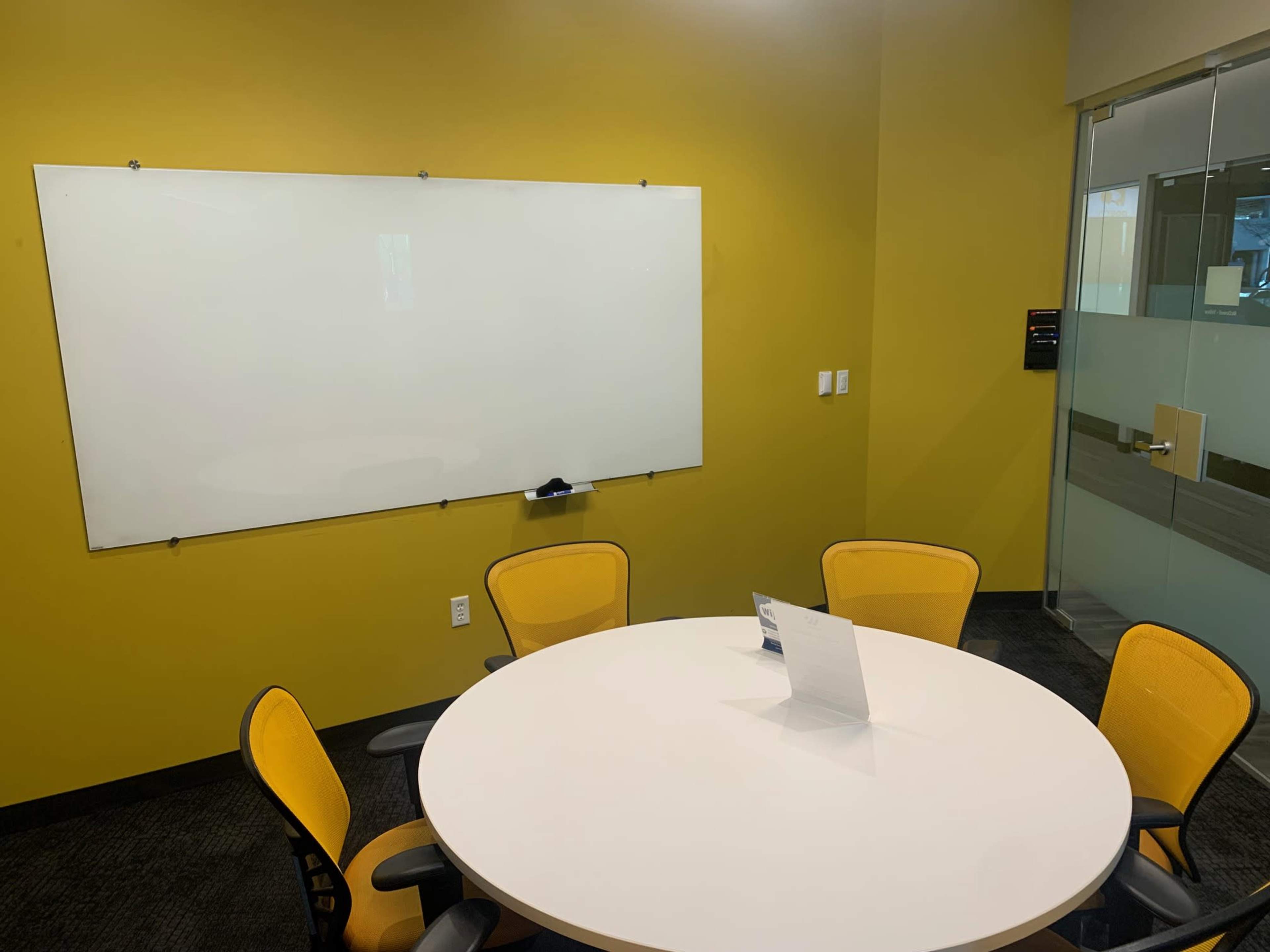 A small meeting room features a whiteboard, a round table with five yellow chairs, and a glass wall.
