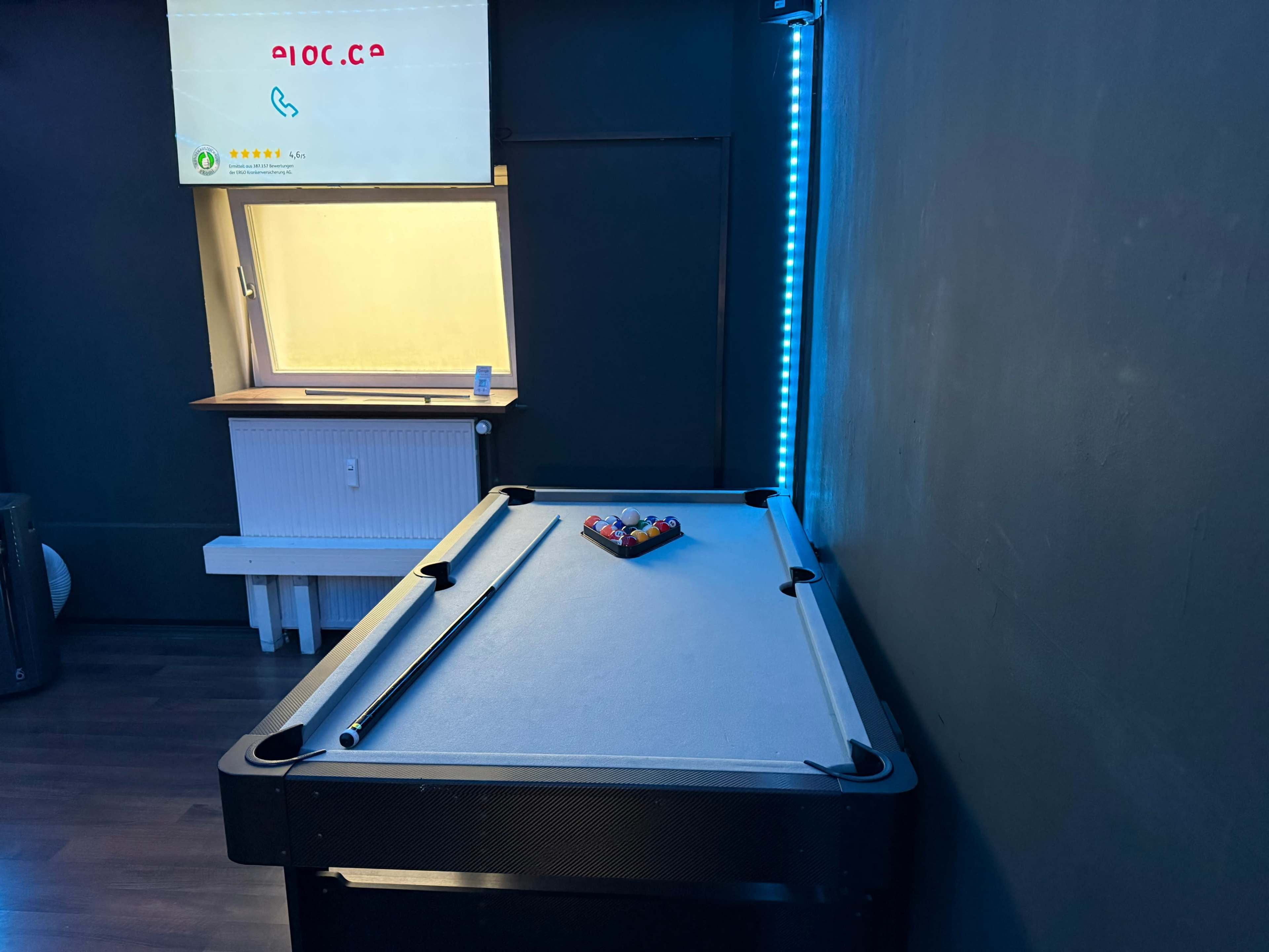 A pool table with colored balls arranged on it is placed in a room with blue LED lighting and a window displaying a yellow tint.