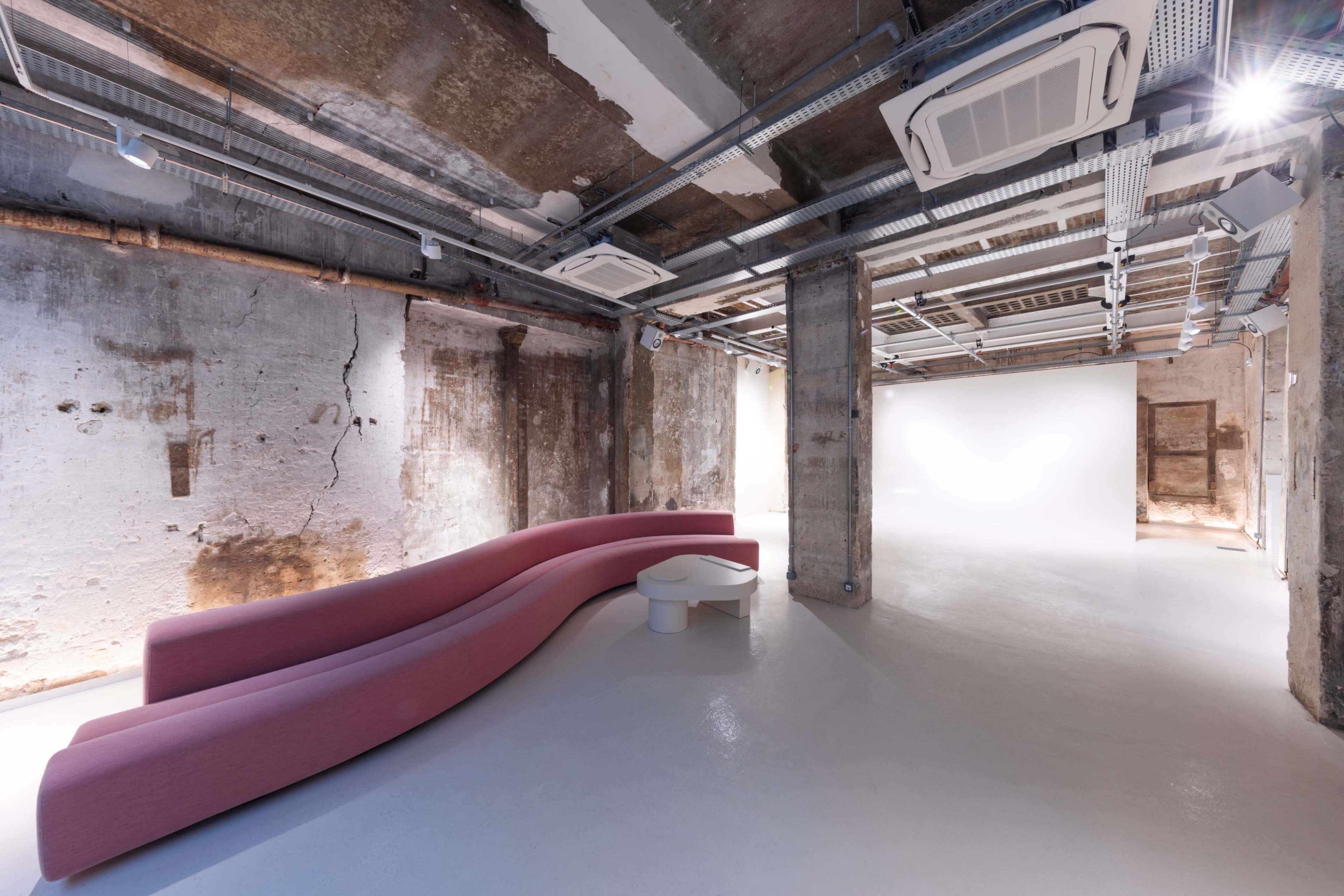 The image shows a modern, minimalist interior space featuring a curved pink sofa and a small white table under industrial-style ceilings.