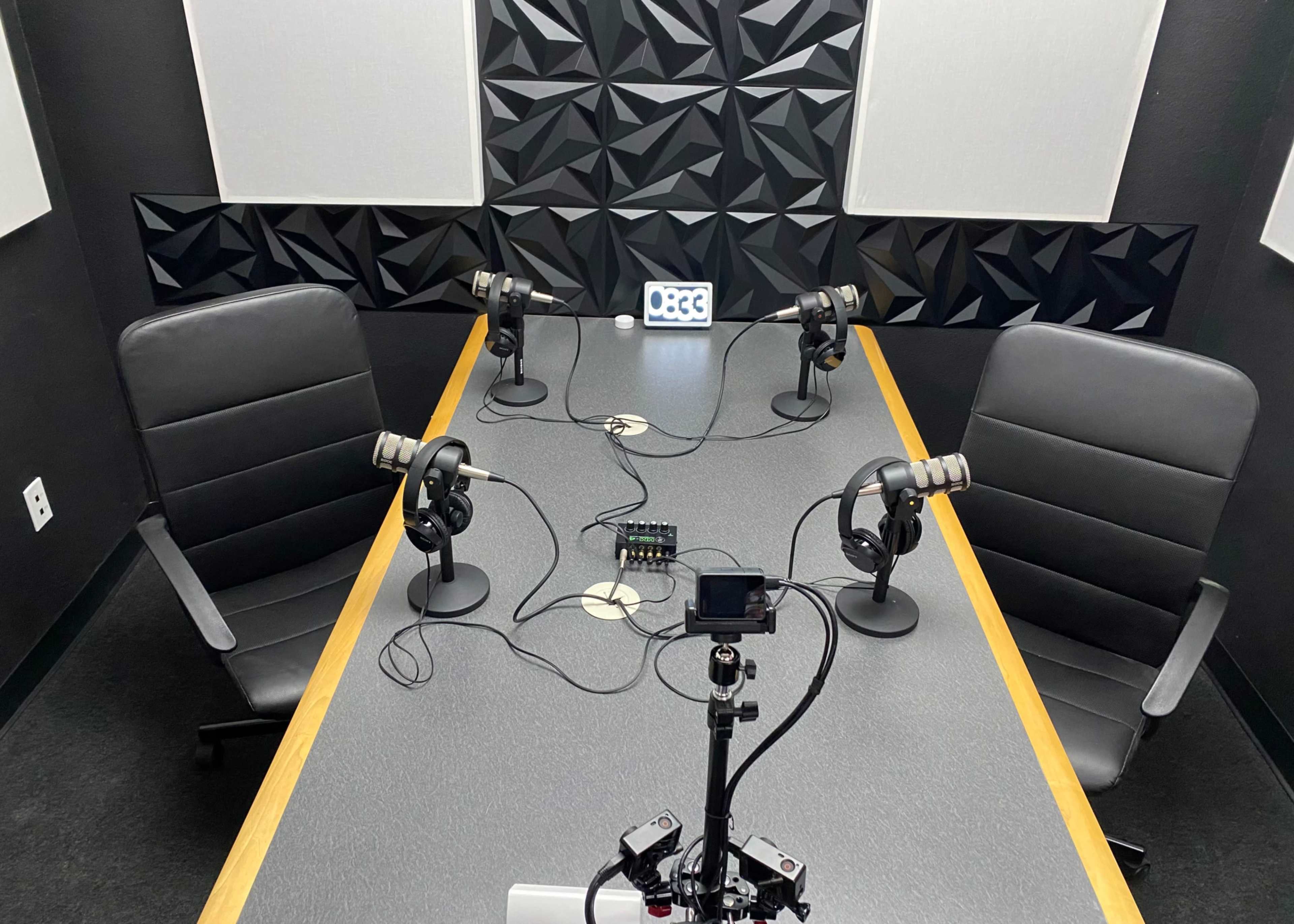 A podcast recording studio features a rectangular table with four microphones, two chairs, and a device for audio mixing, set against a textured black wall.