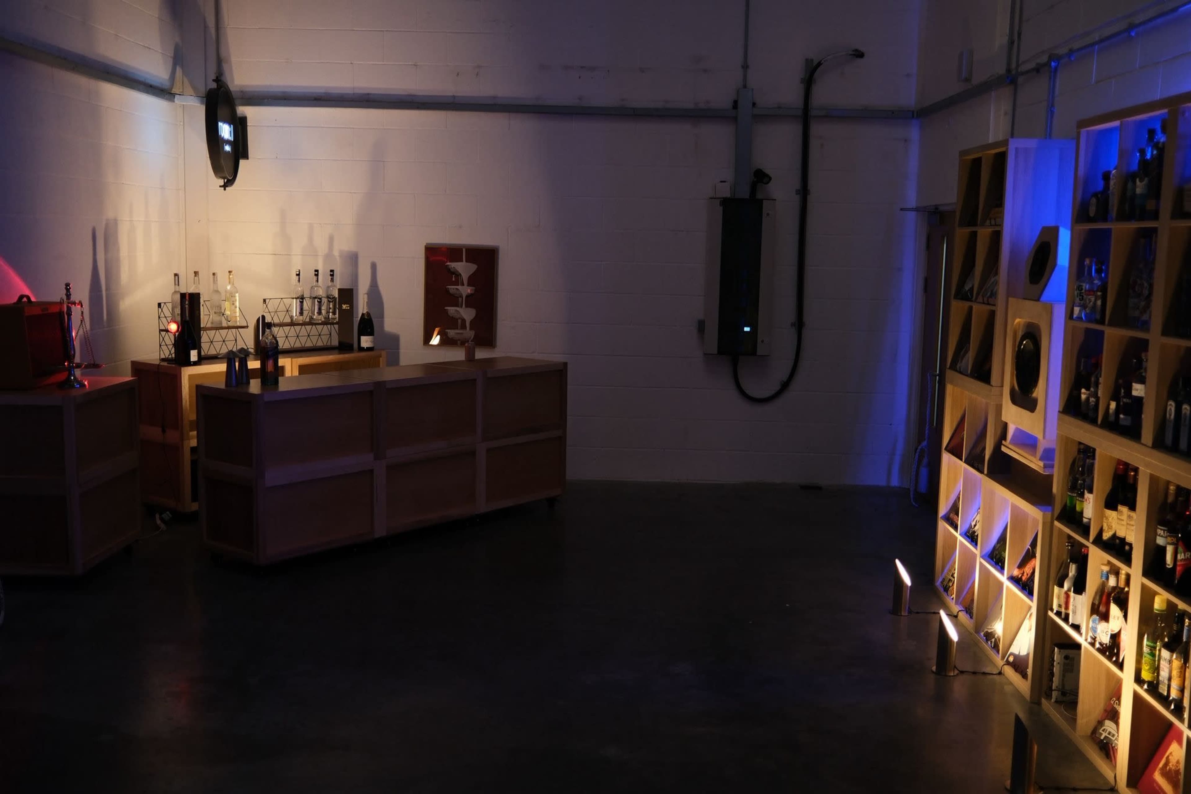 The image shows a minimalist bar area with two wooden counters, shelves containing various bottles, and low lighting in a concrete space.