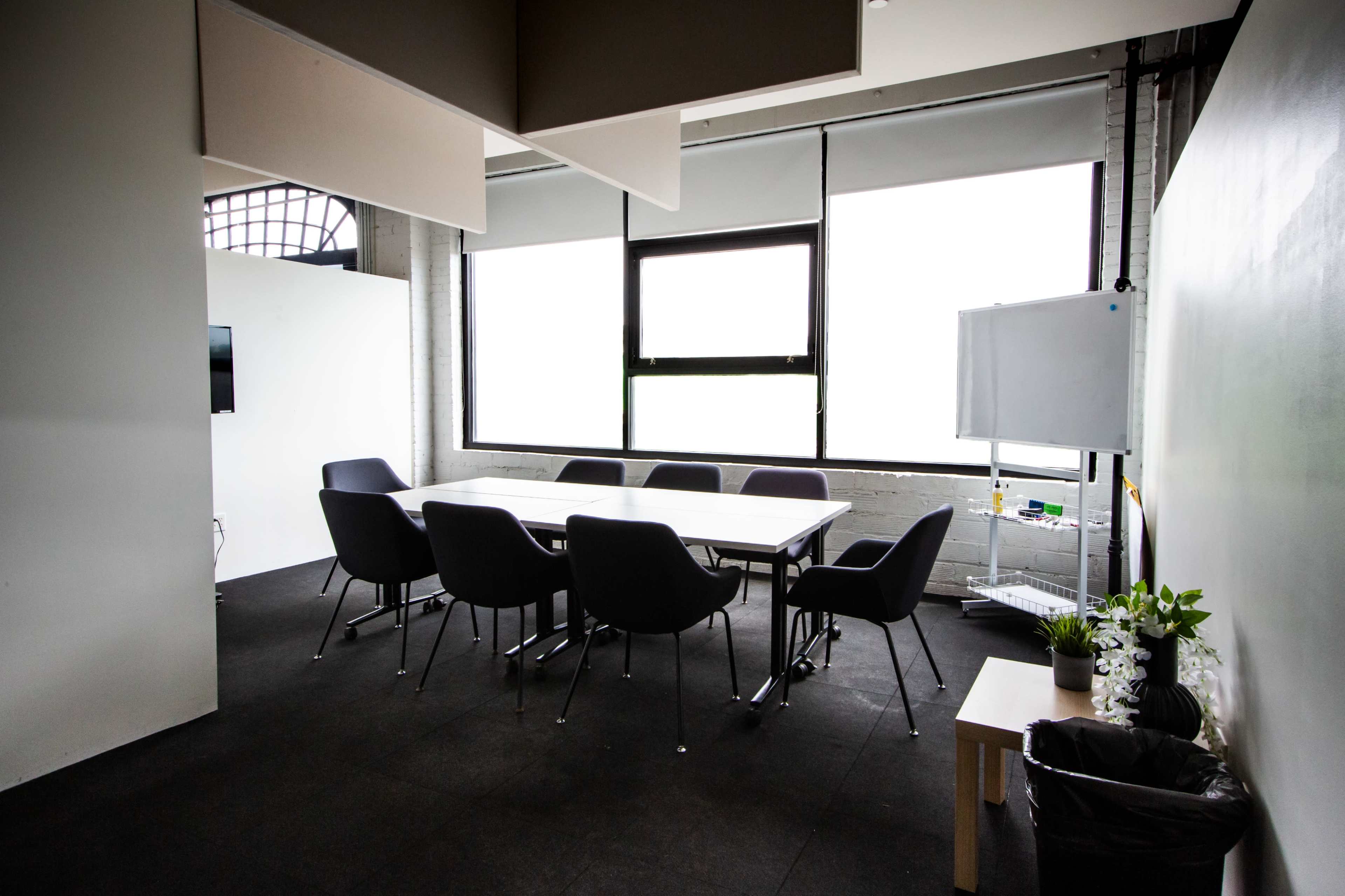 A modern conference room features a large table surrounded by chairs, with windows providing natural light and a whiteboard in the background.