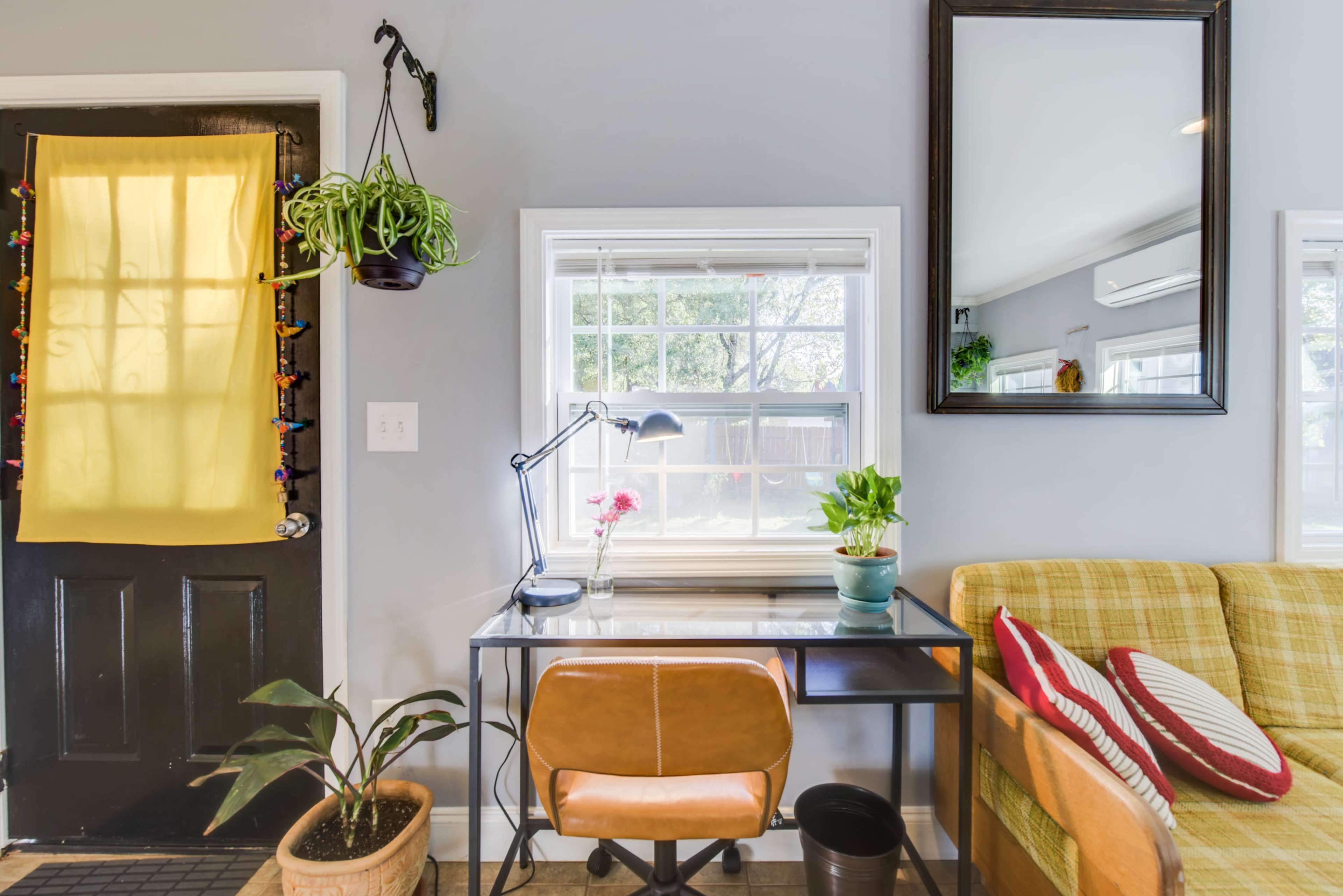 A small home office setup features a desk with a lamp and potted plants, adjacent to a yellow curtain over a door and a couch in a cozy room.