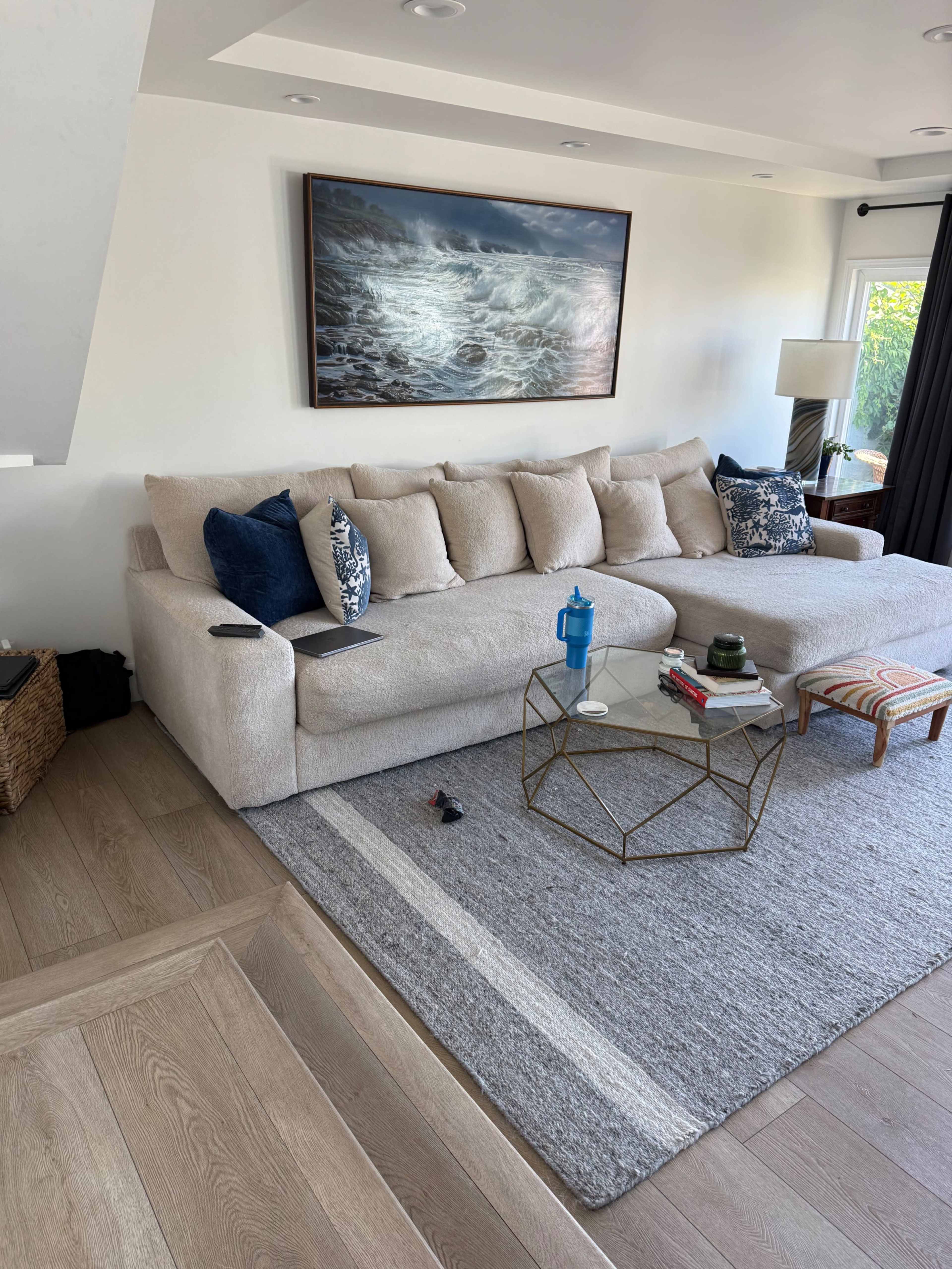 The image shows a living room with a large beige sectional sofa, a coffee table with various items, a patterned chair, and a large painting of waves on the wall.
