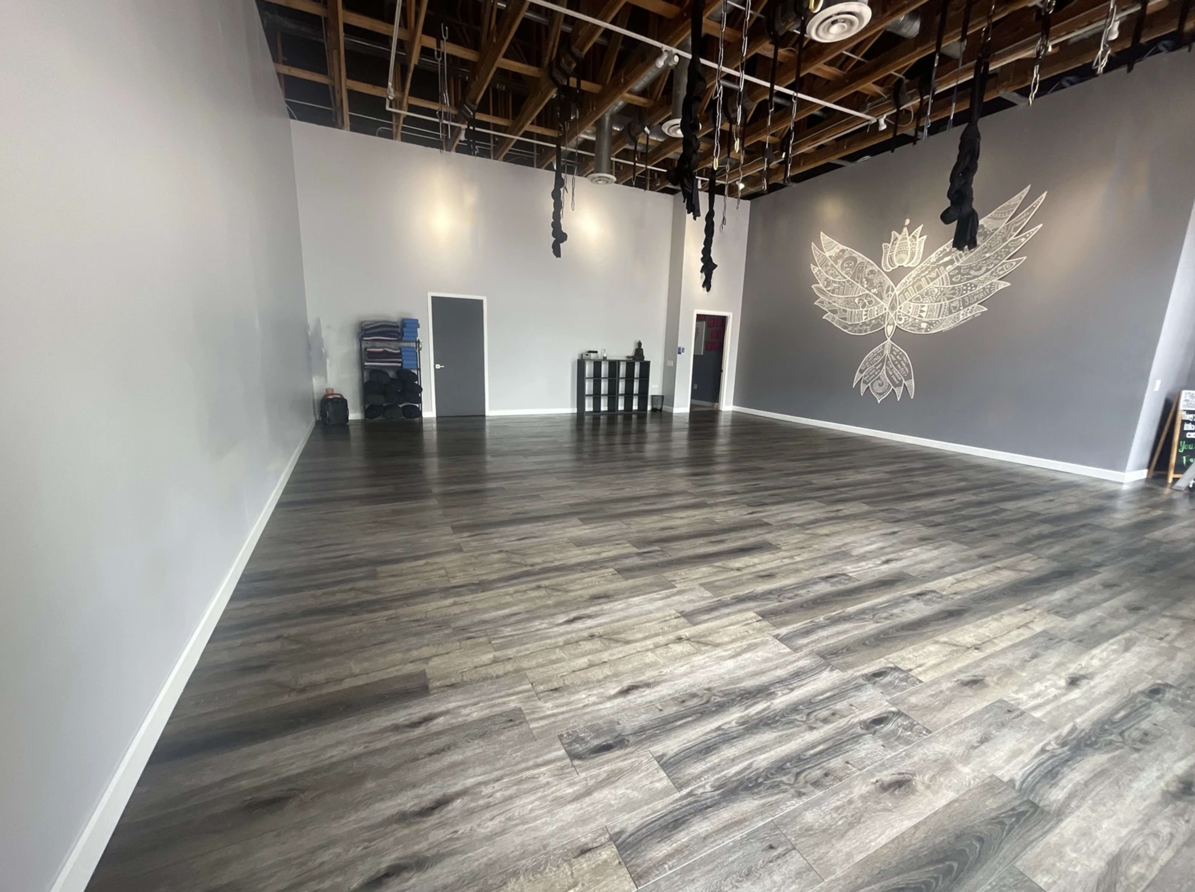 A spacious, empty studio with wooden flooring, white walls, and a gray accent wall featuring a large design, along with some equipment in the corner.
