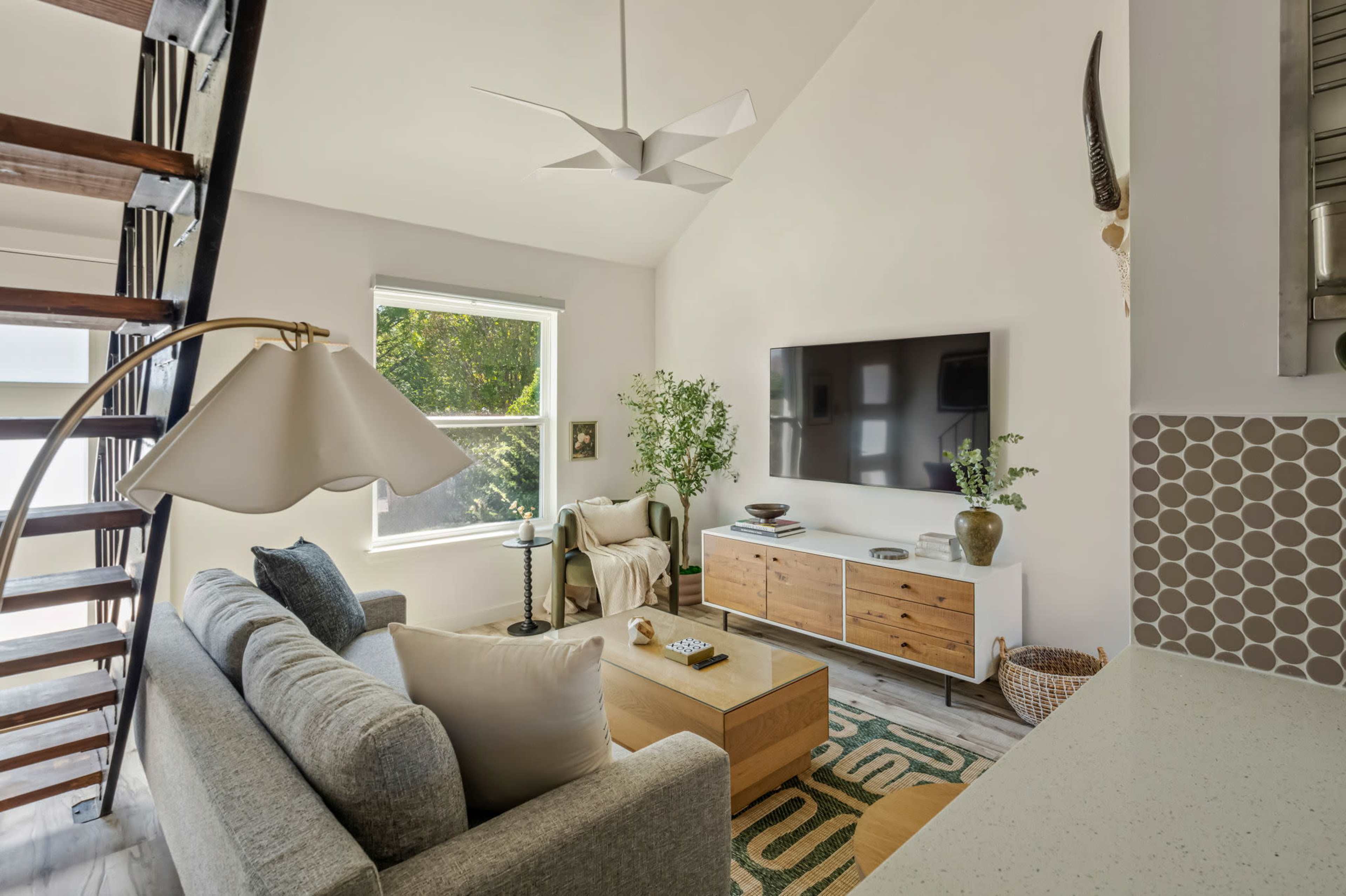 The image shows a modern living room with a large window, a cozy sofa, a wooden coffee table, and a mounted television.