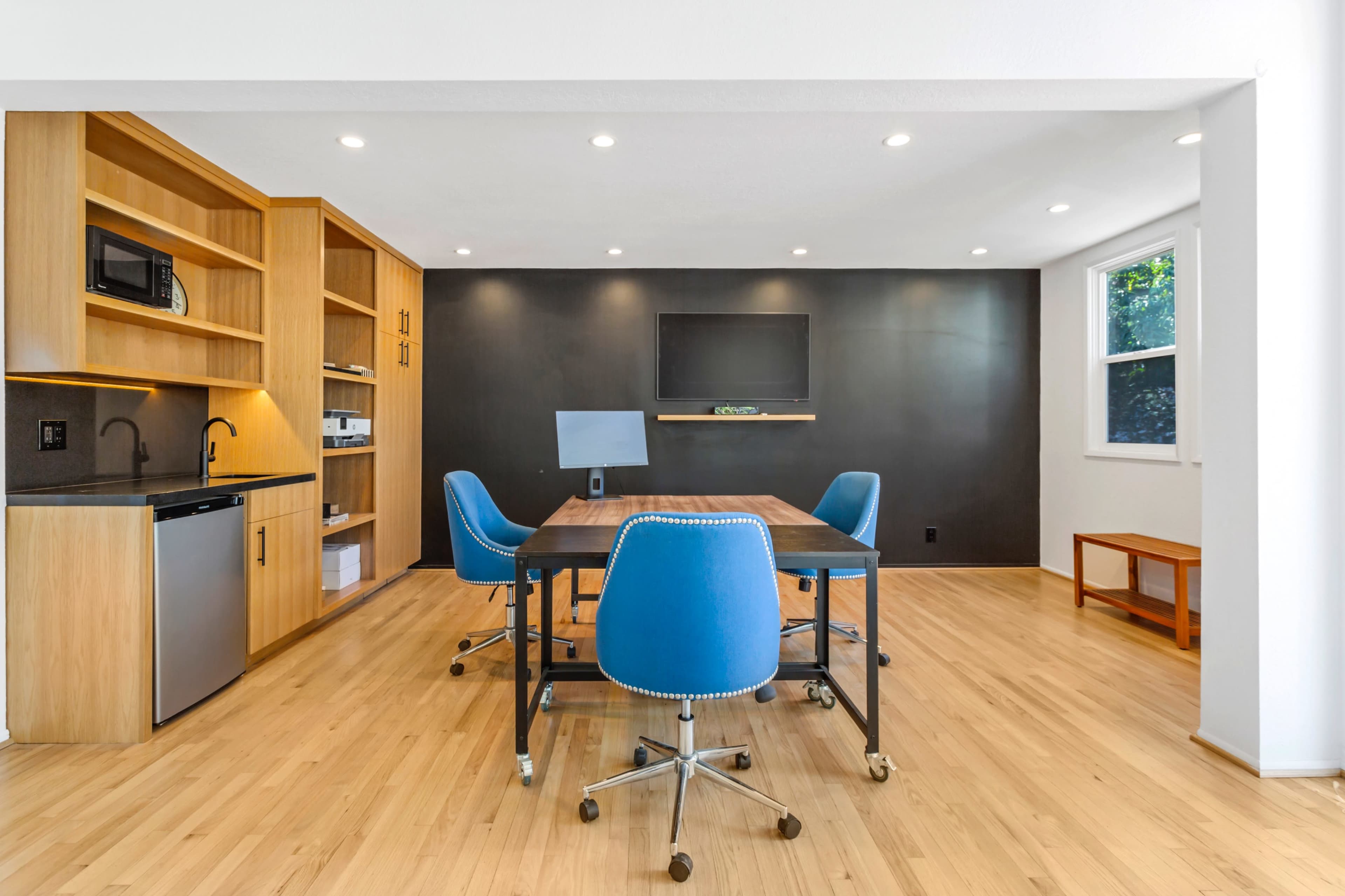 A modern office space features a wooden table surrounded by blue chairs, with a kitchenette and a wall-mounted television in the background.