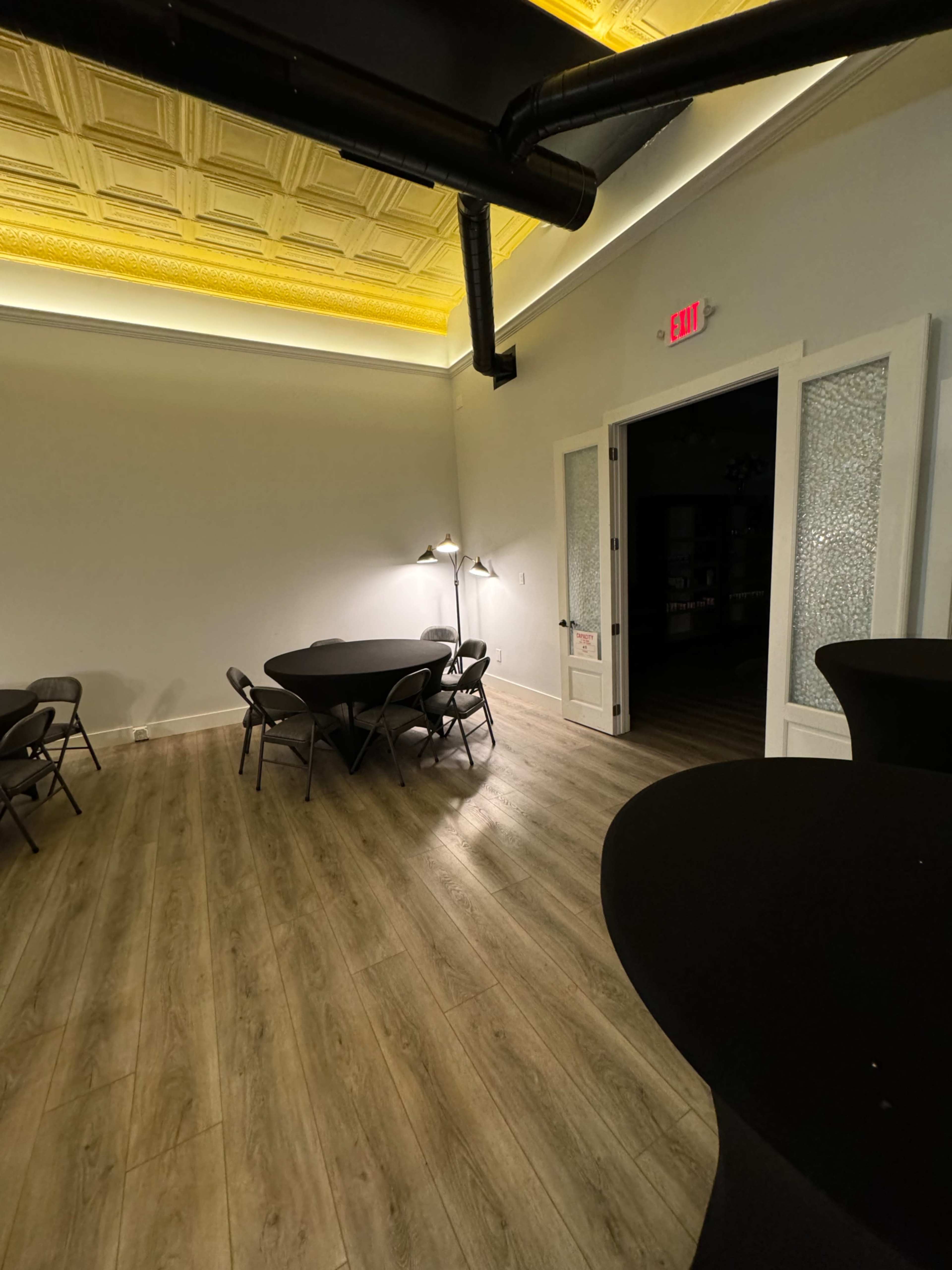 A room with a light-colored ceiling and decorative molding features several black folding tables and chairs, with an exit door on one side.