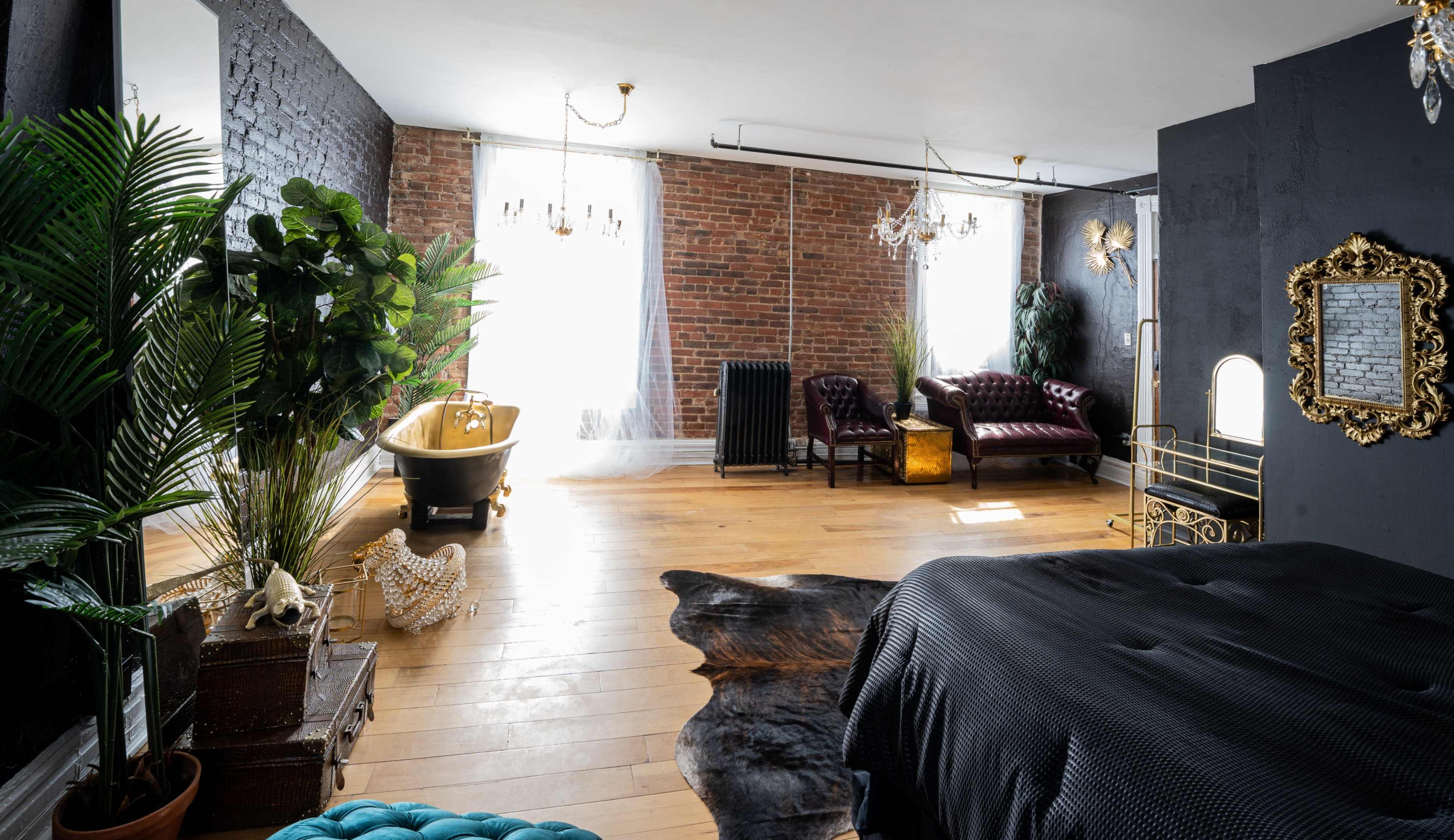 The image shows a spacious bedroom featuring exposed brick walls, a vintage clawfoot bathtub, a leather seating area, and large windows letting in natural light.
