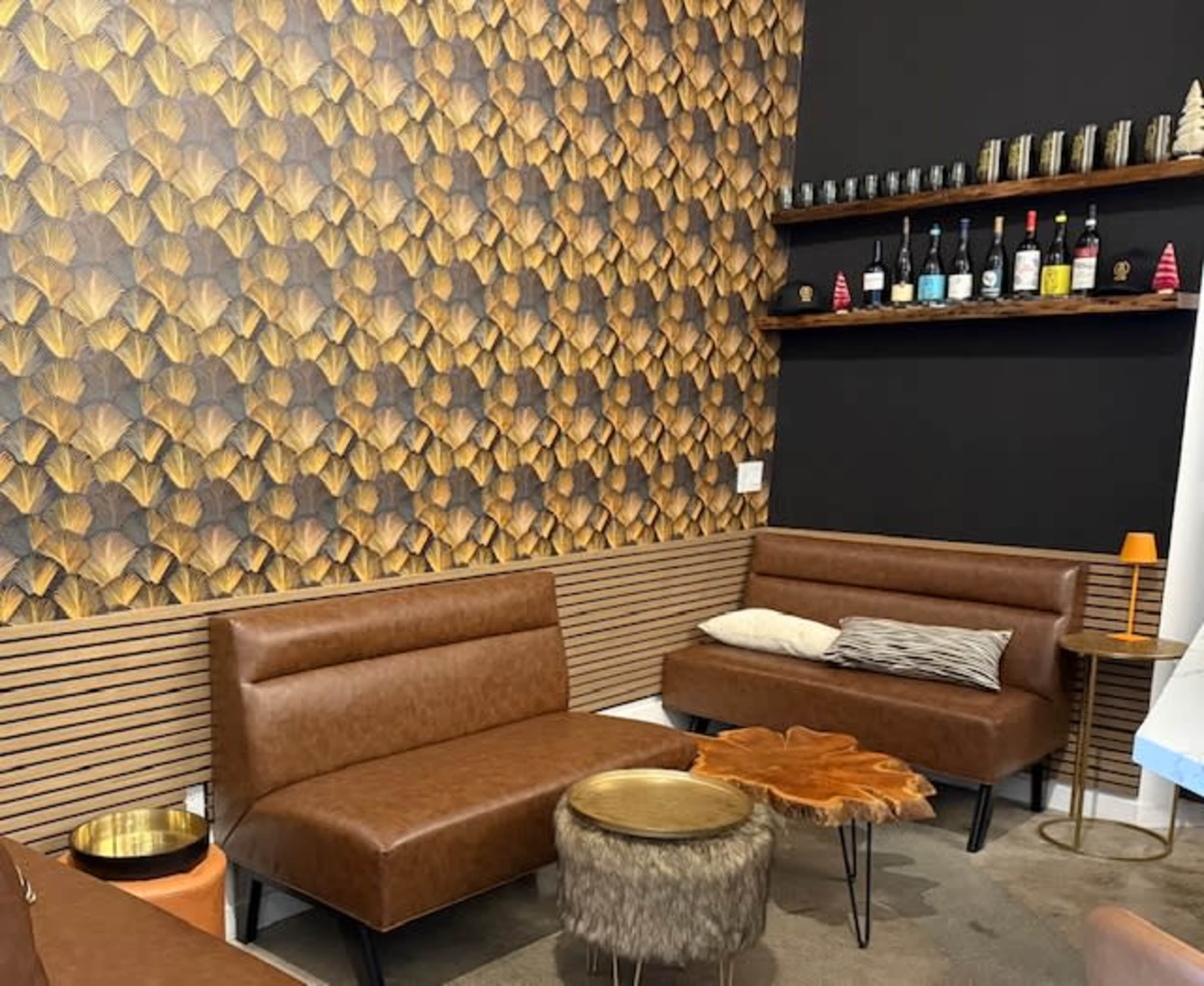 The image shows a cozy seating area with brown leather couches, a decorative patterned wall, and a shelf displaying various bottles.