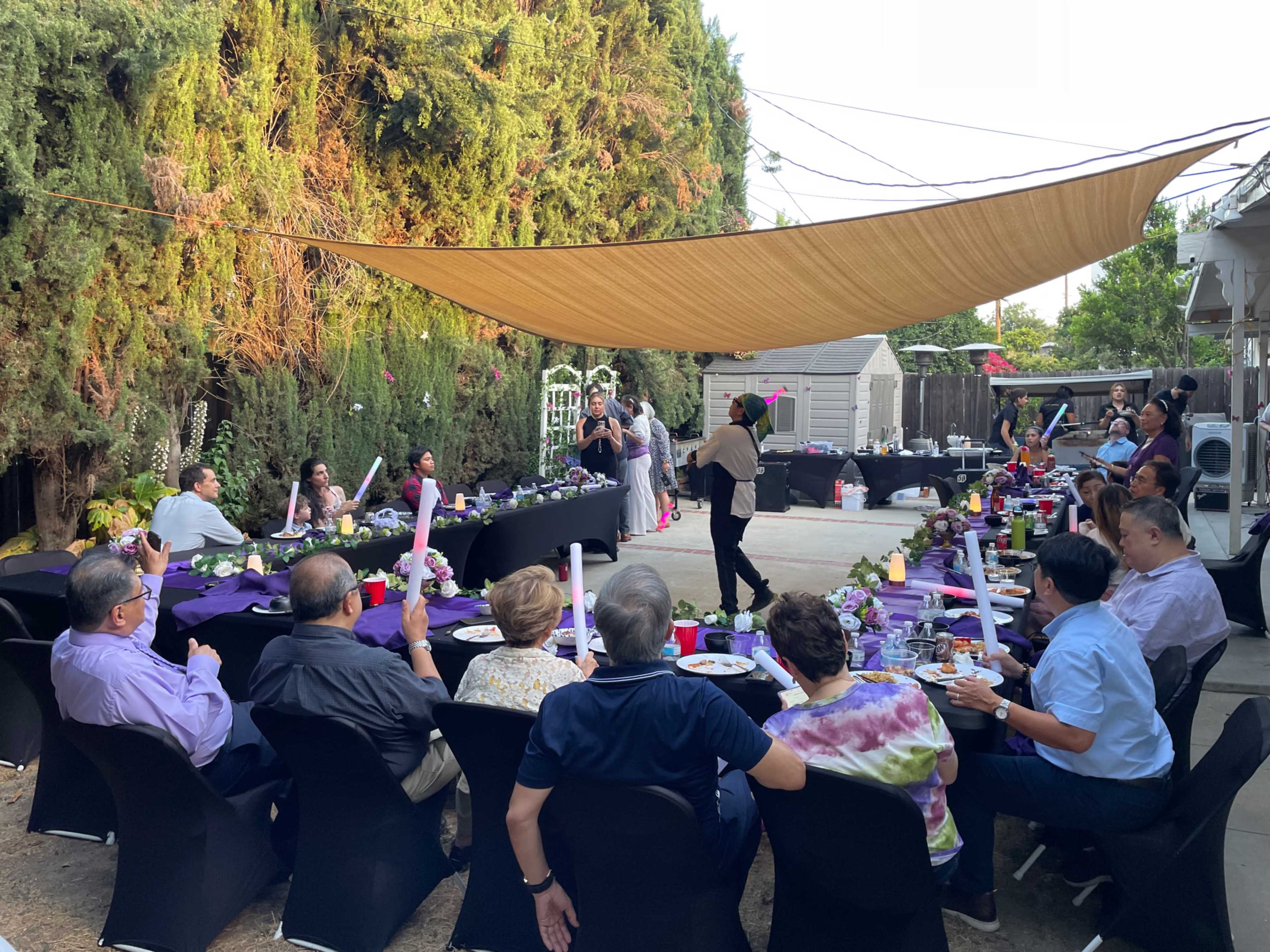 A group of people is seated at a long table under a shaded canopy in a backyard, enjoying a meal surrounded by greenery.
