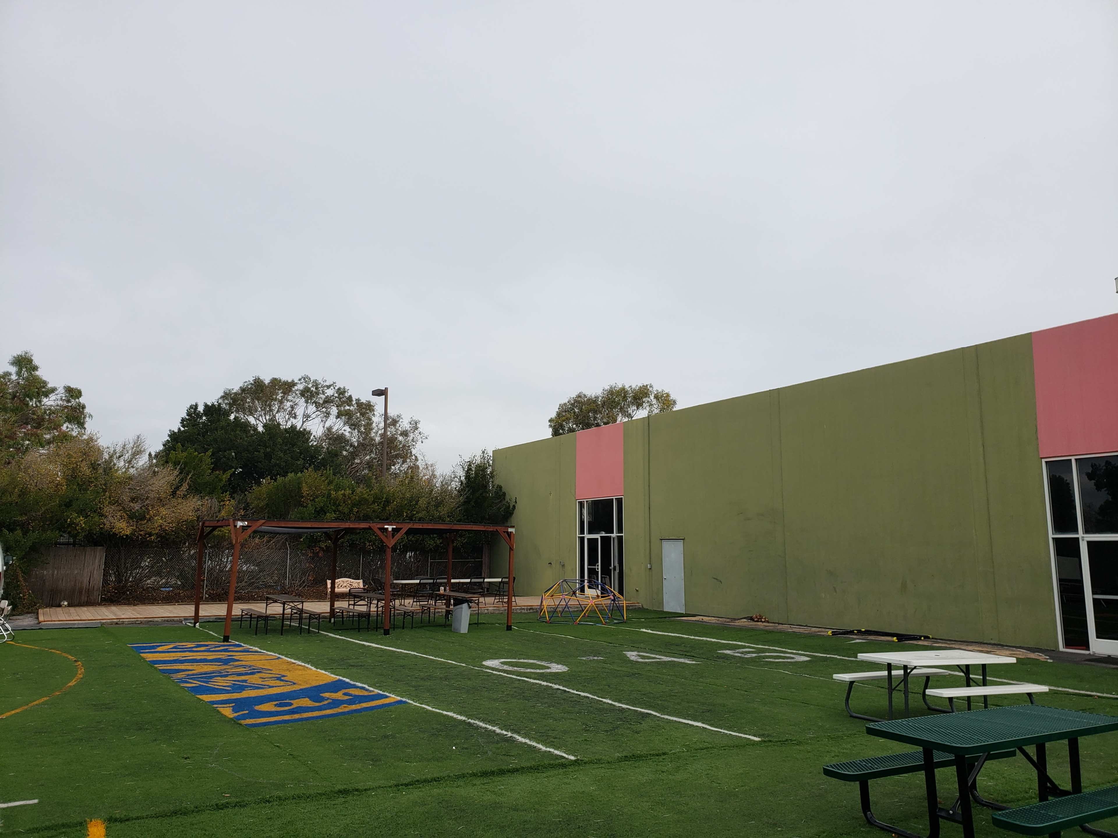 A grassy outdoor area with picnic tables, a wooden structure for shade, and a small playground beside a colorful building.