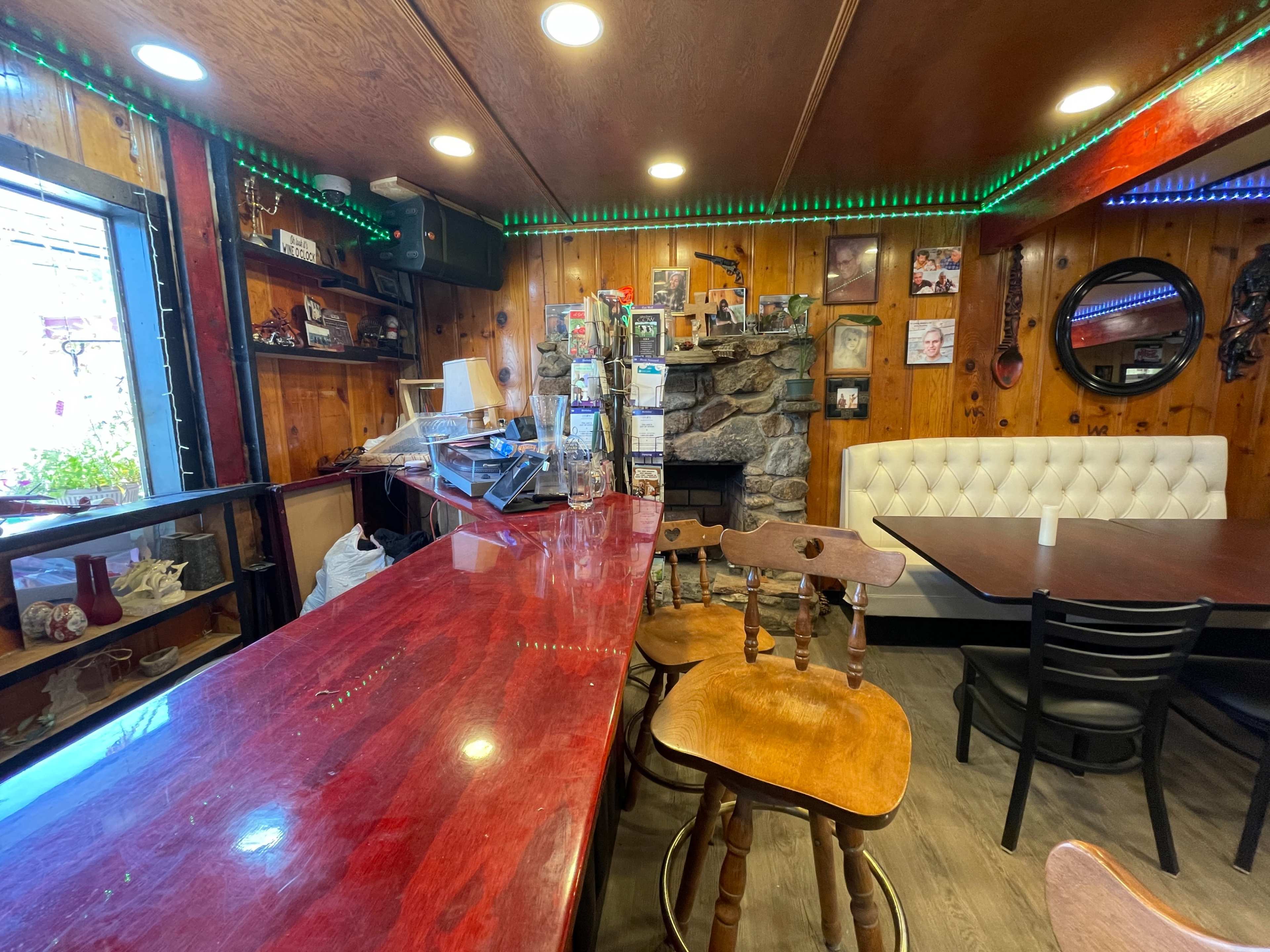 A wooden bar area with a polished counter, surrounded by tables and decorated walls, including a stone fireplace.