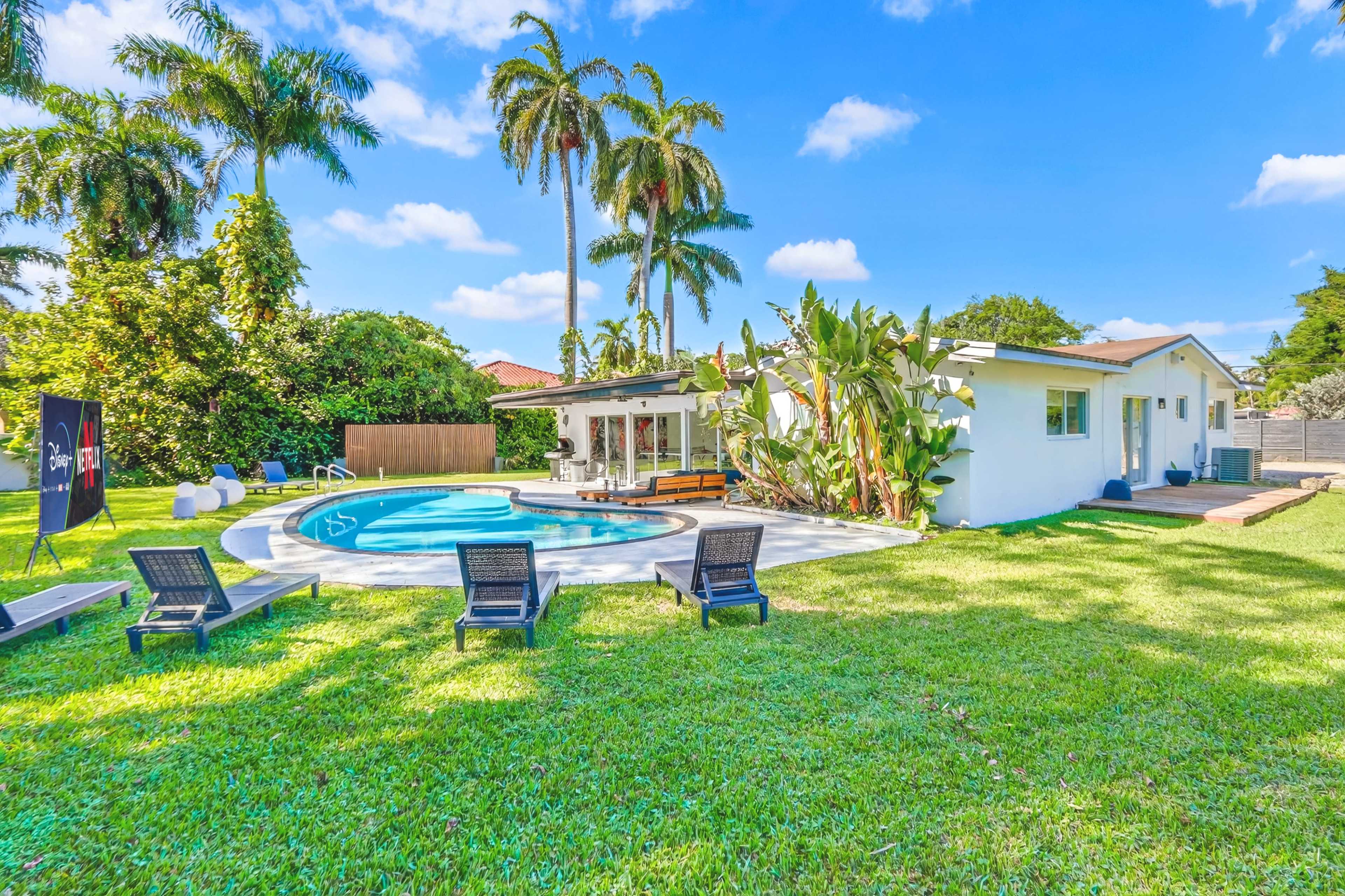 The image shows a backyard with a swimming pool, surrounded by palm trees and a grassy area, with lounge chairs positioned nearby.
