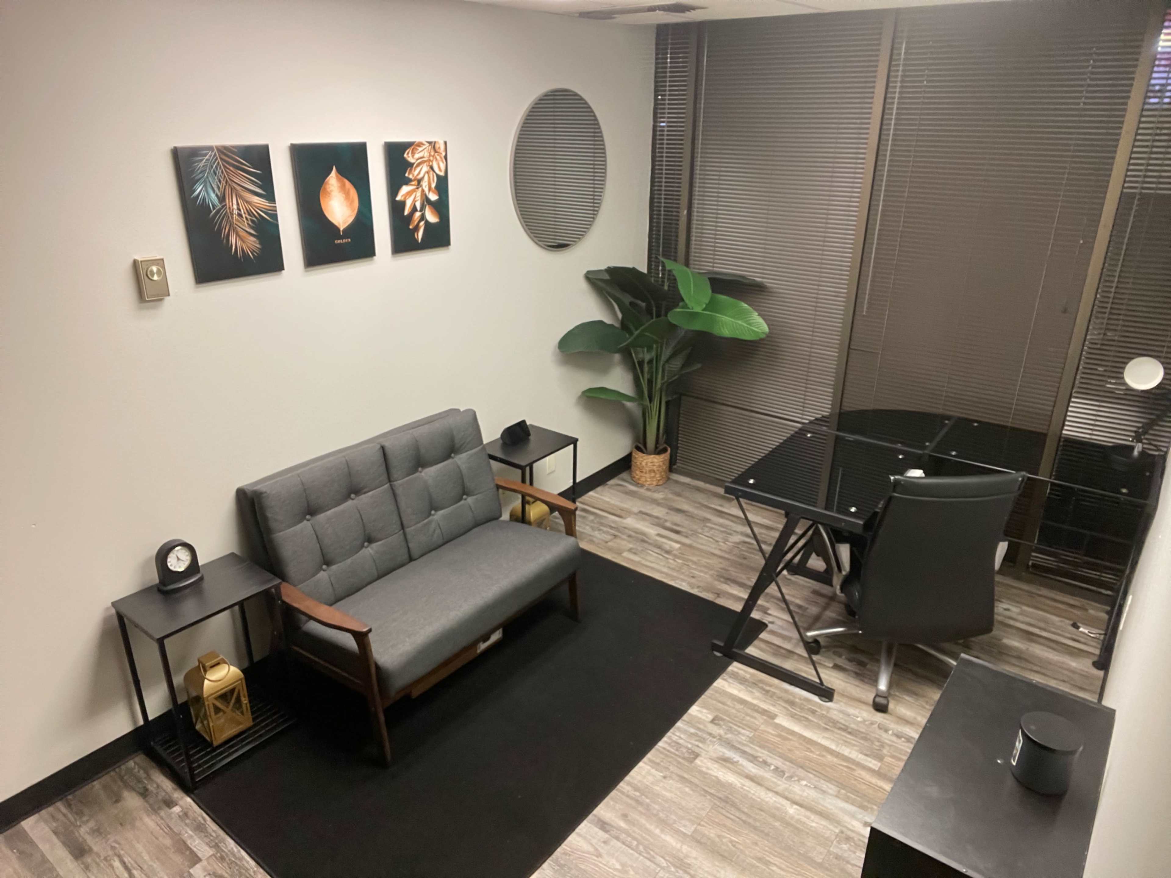 The image shows a small office space featuring a gray sofa, a glass desk, and a potted plant, complemented by framed artwork on the wall.
