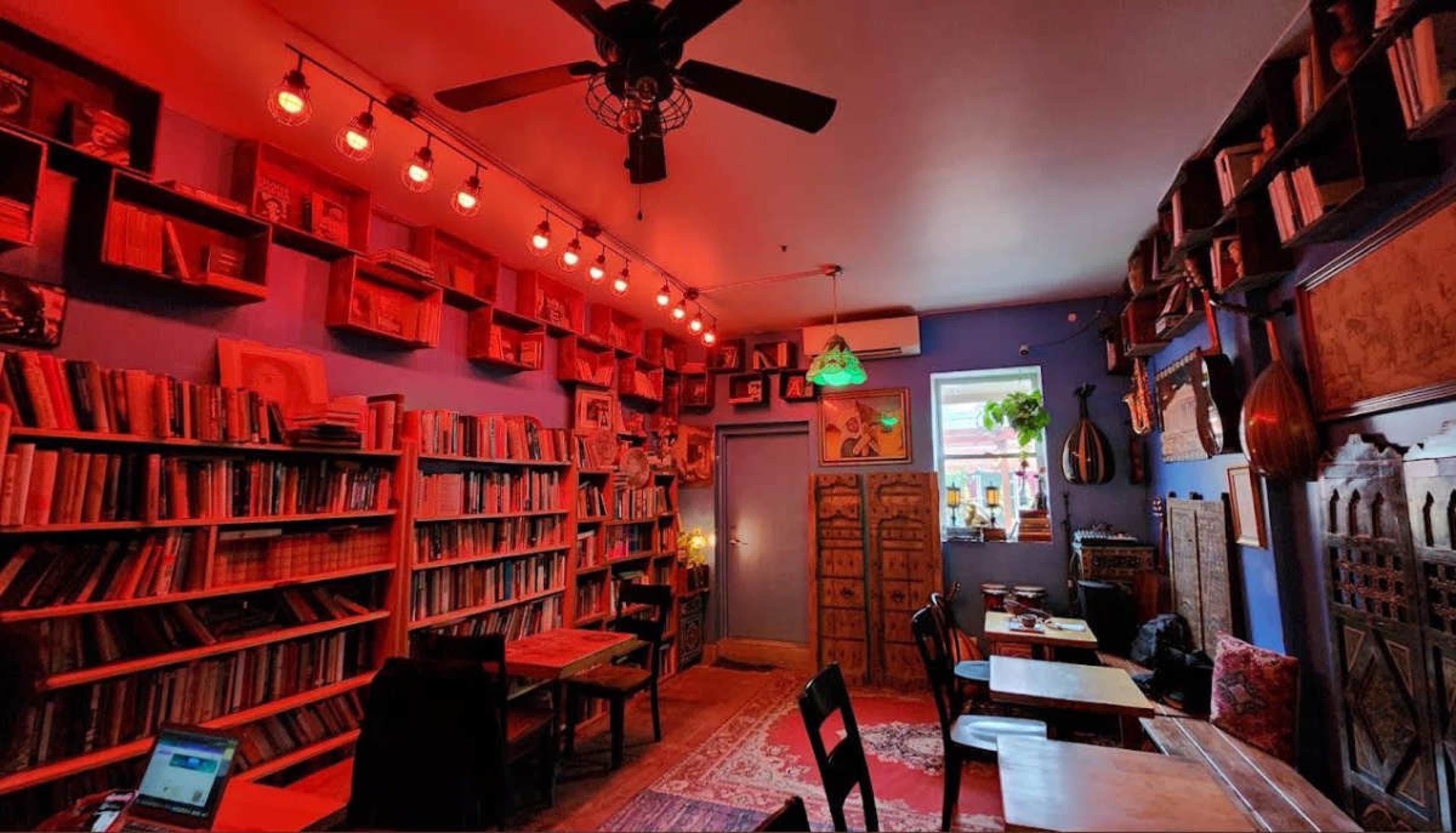 The image depicts a cozy bookshop with red accent lighting, filled with bookshelves lining the walls and wooden tables scattered throughout the space.