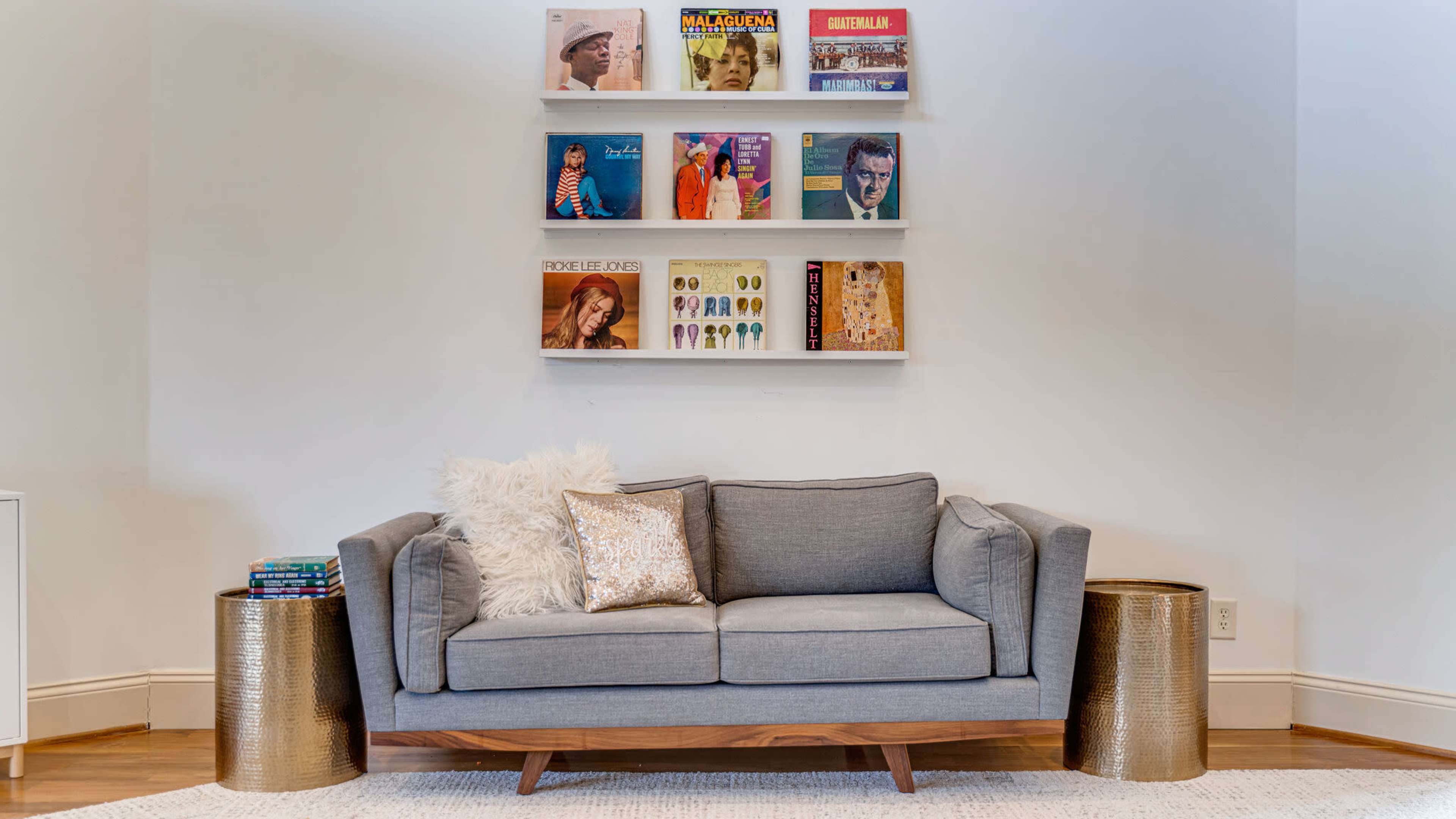 A gray sofa with a decorative pillow is positioned in front of a wall-mounted shelf displaying colorful vinyl records, flanked by two gold end tables.