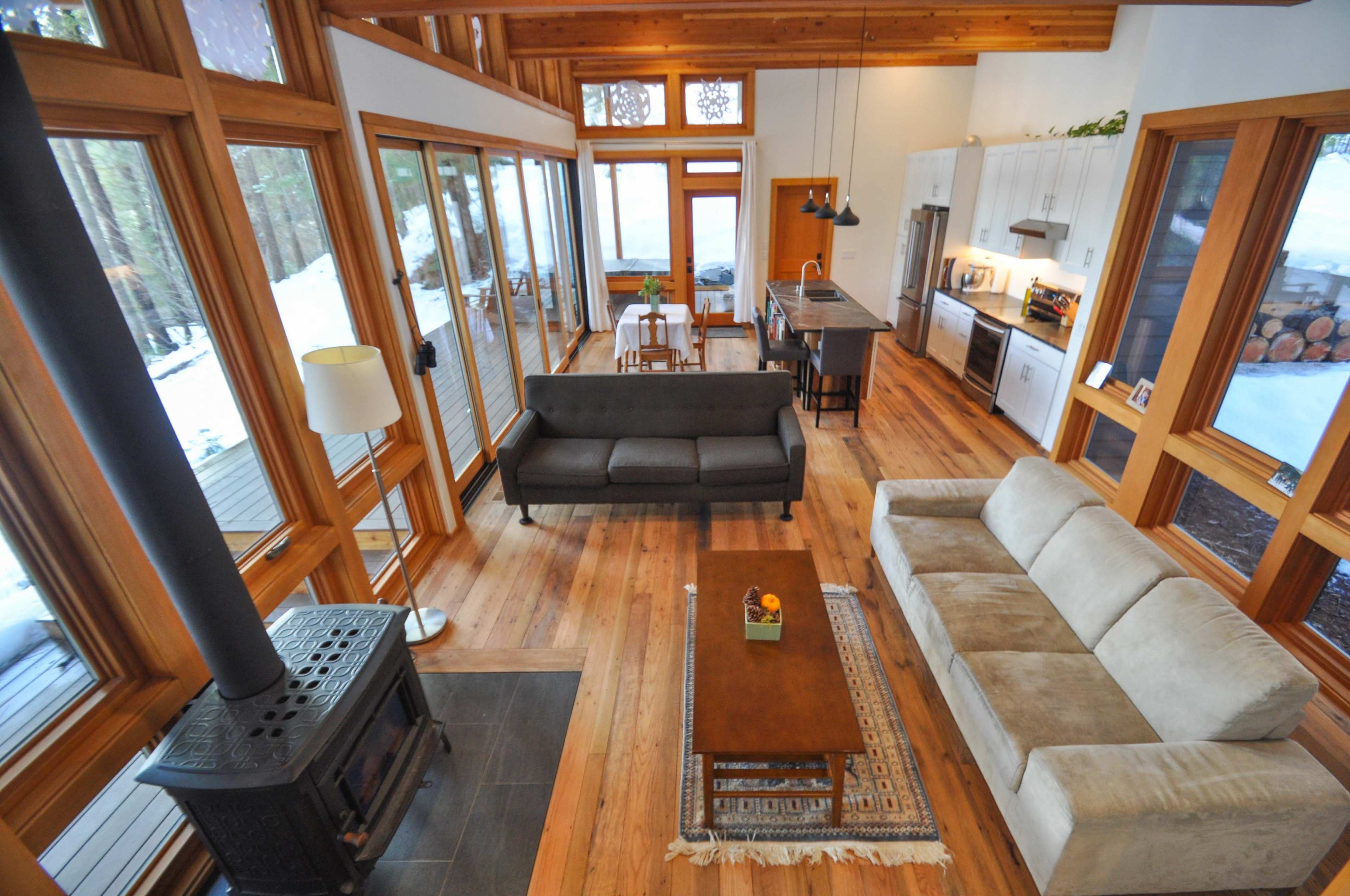 A spacious, modern living room features large windows, wooden beams, and a mix of seating arrangements with a kitchen area visible in the background.