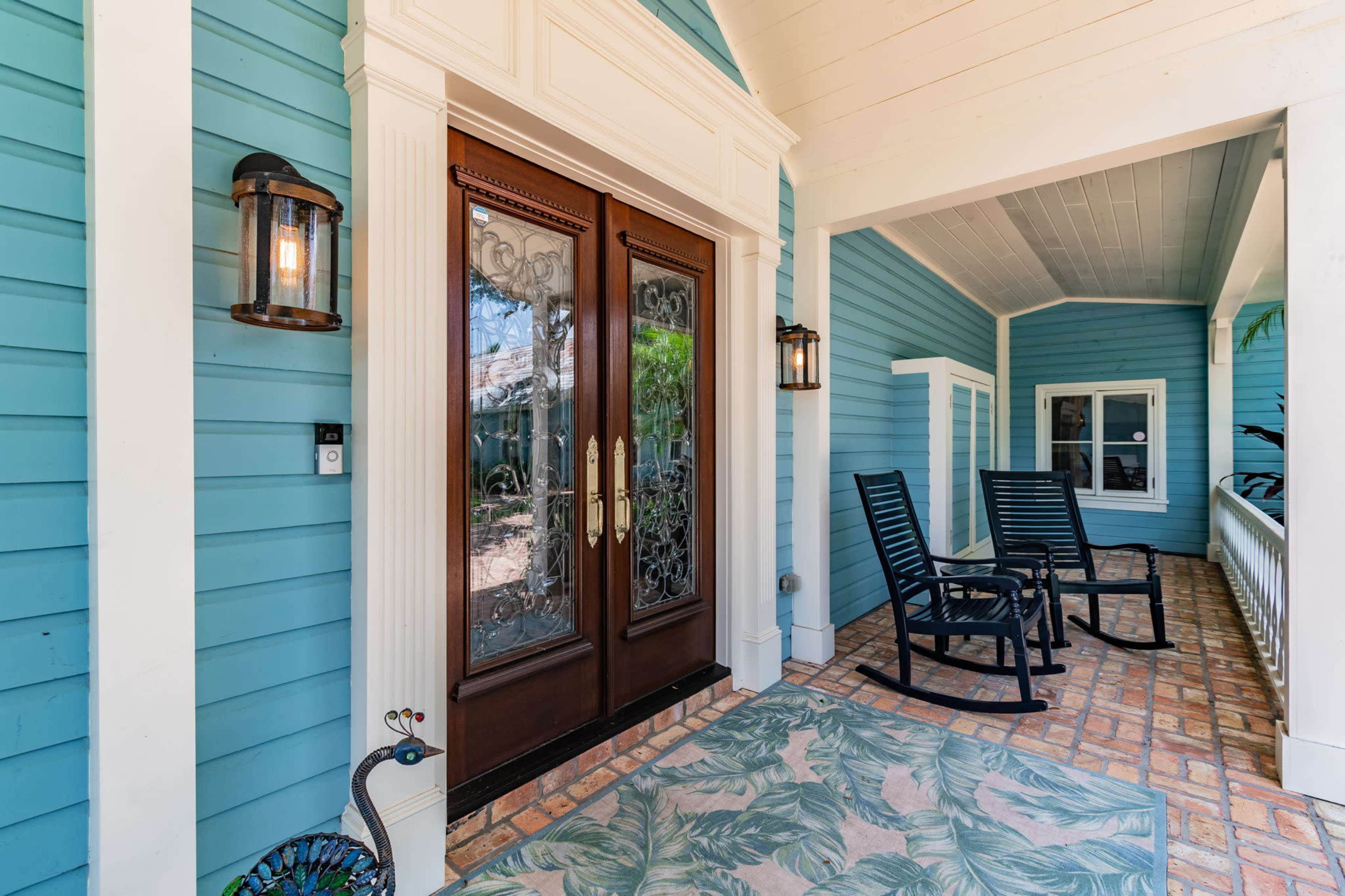 A front porch with blue wooden siding, featuring double doors with decorative glass, a patterned rug, and two black rocking chairs.