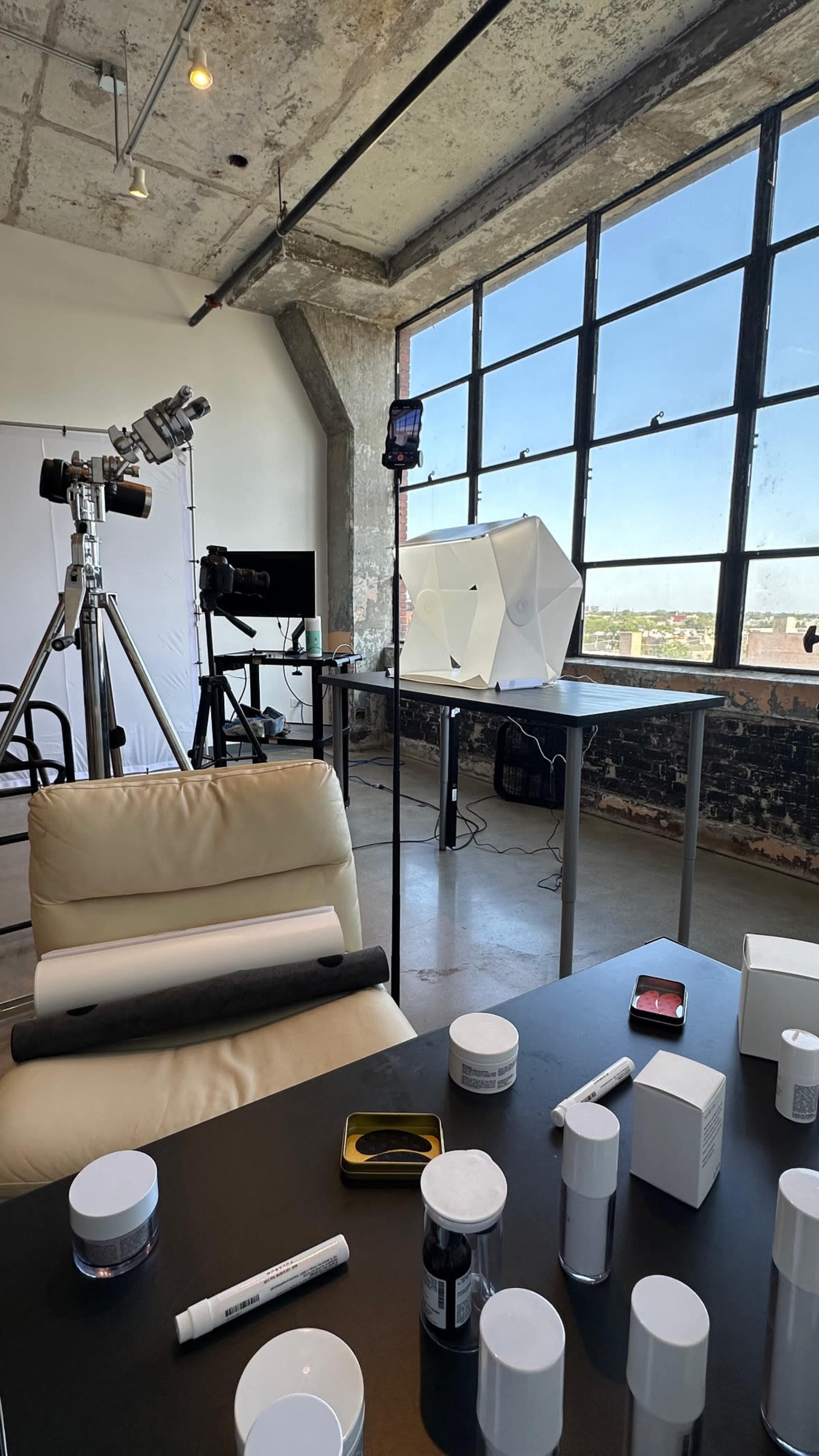 The image shows a bright studio space with large windows, equipped with photography equipment, a table with various cosmetic containers, and a backdrop setup.