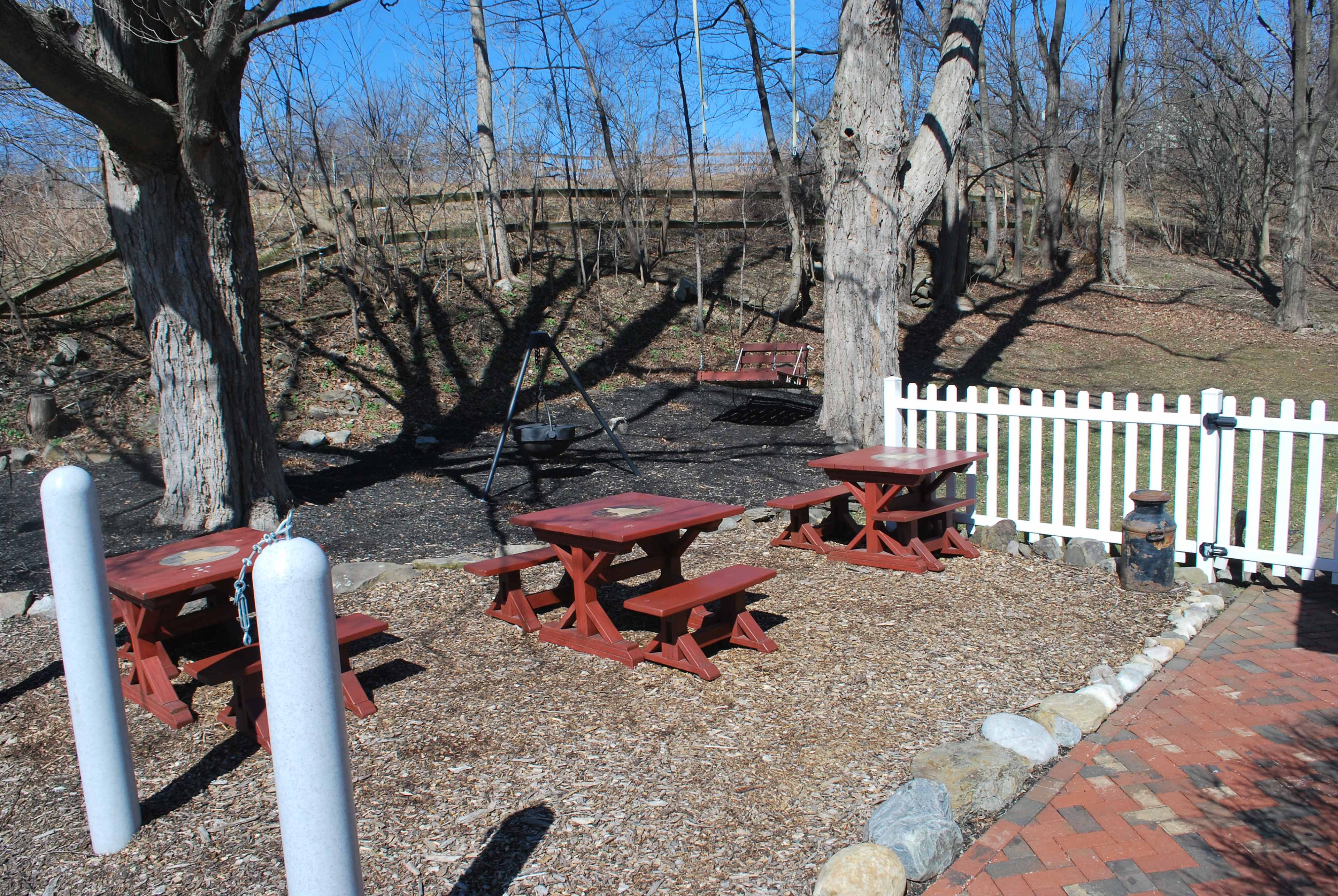 A rustic outdoor seating area with wooden picnic tables surrounded by trees and a white picket fence.
