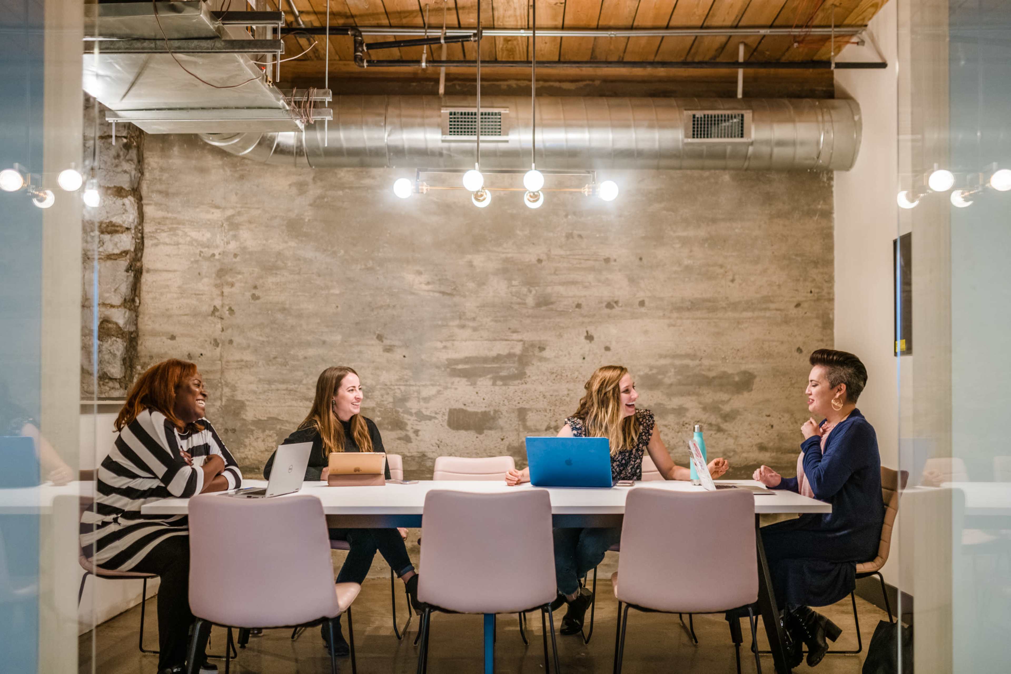 A group of five women is seated around a table in a modern meeting room with a concrete wall and overhead lighting.