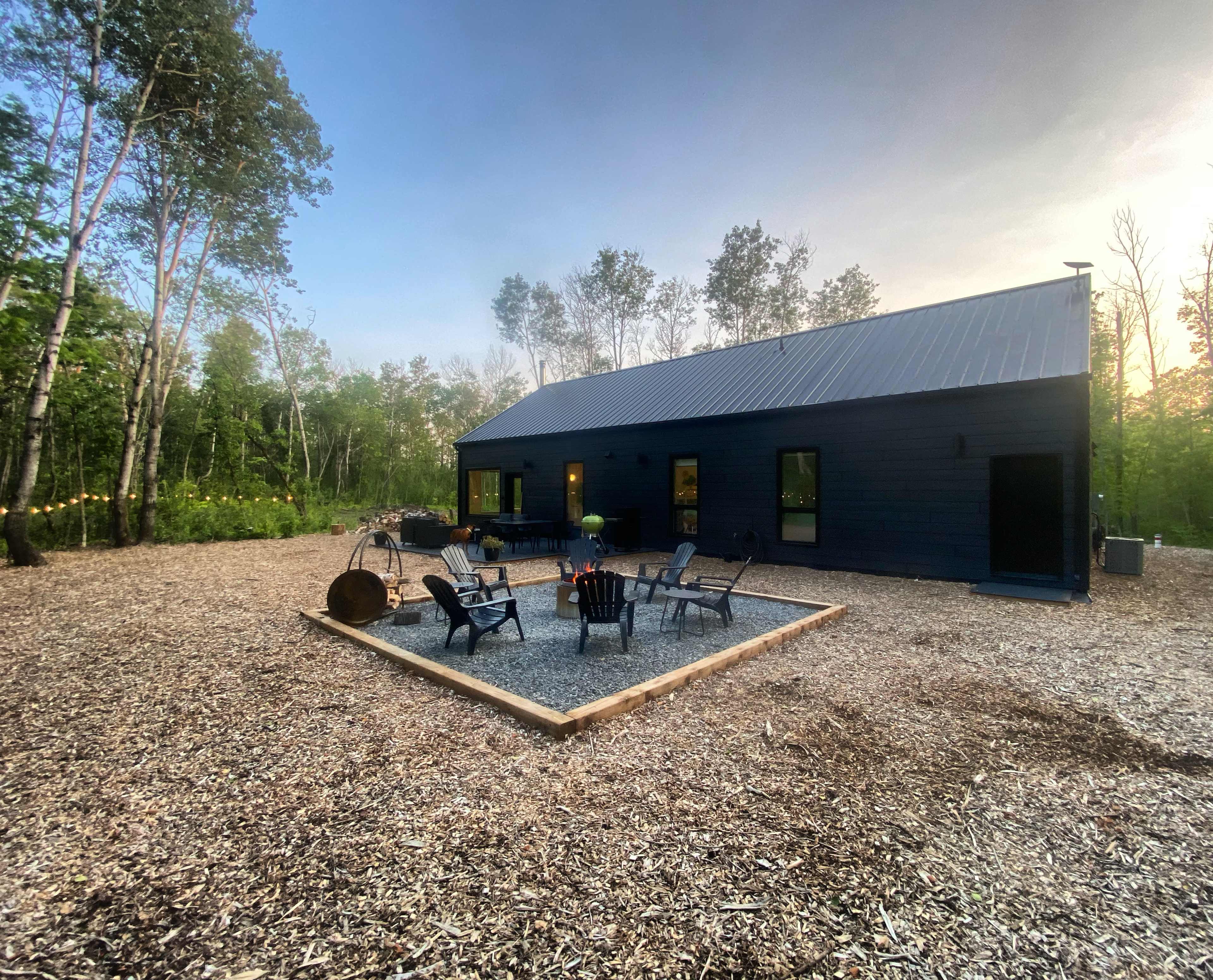 A modern black house with a metal roof is surrounded by a gravel fire pit area and trees.