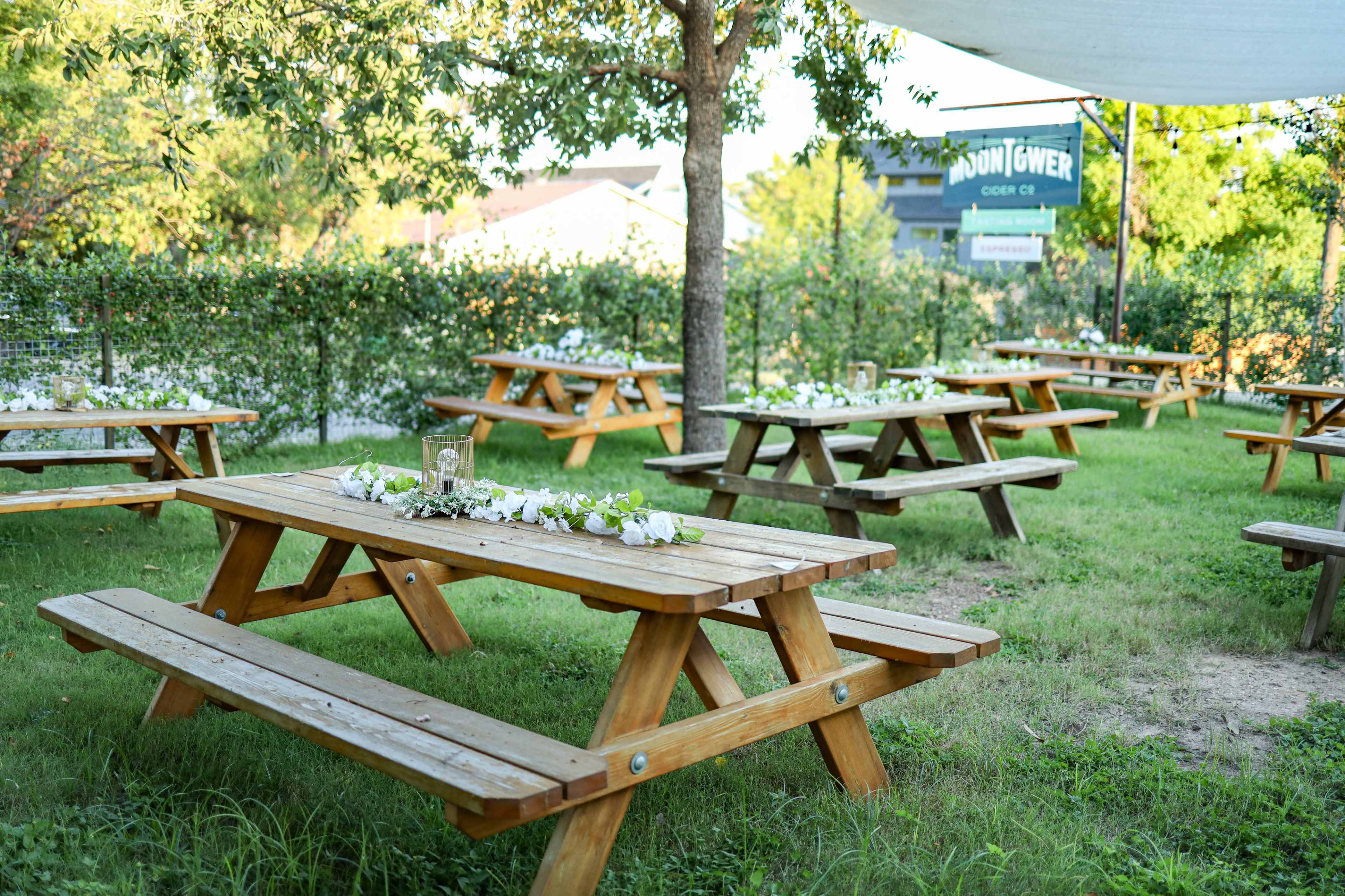 A spacious outdoor area with wooden picnic tables arranged on green grass, adorned with centerpieces of flowers.