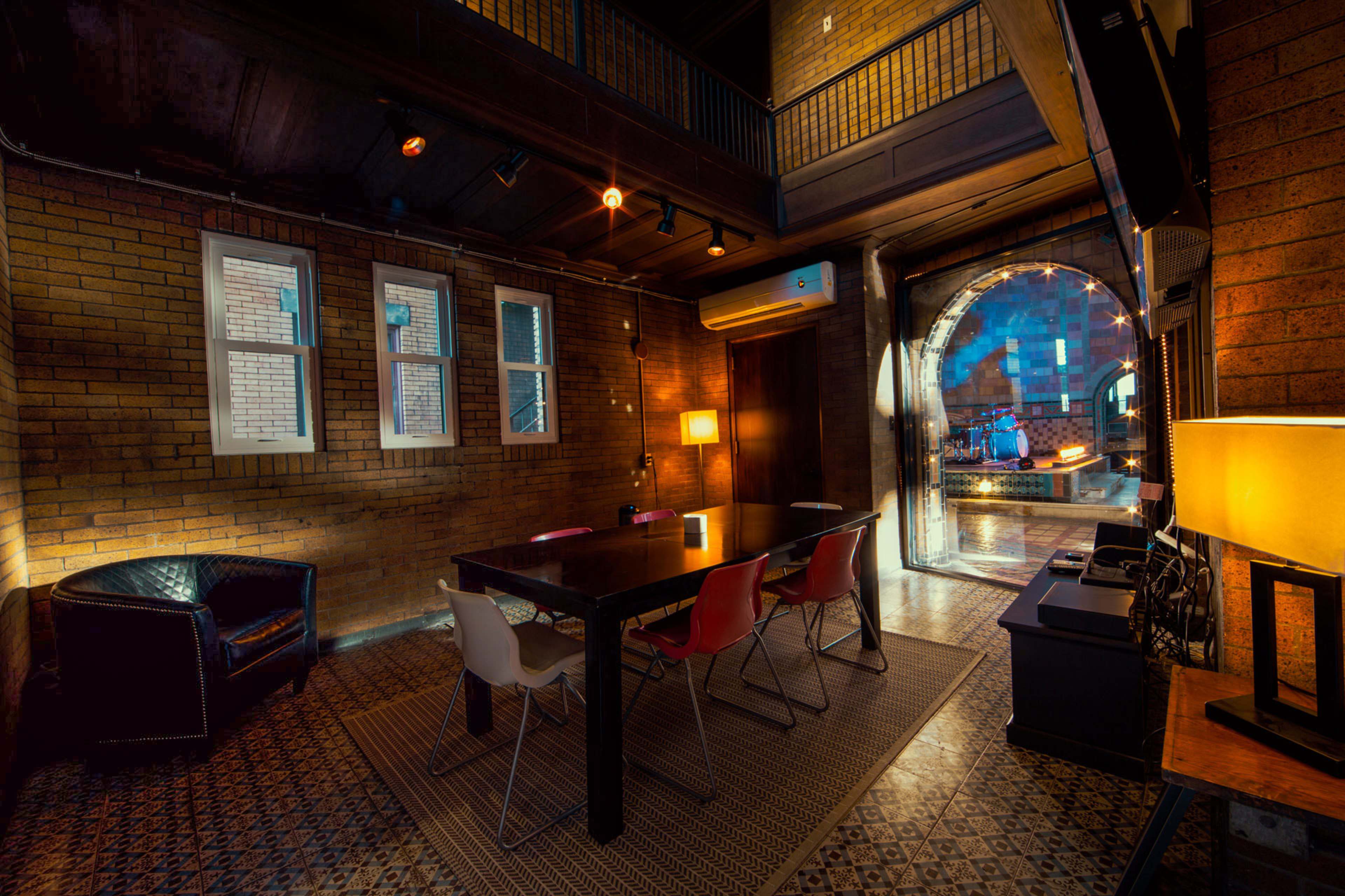 A dimly lit room with exposed brick walls, featuring a large wooden table surrounded by colorful chairs, and an archway leading to another space.