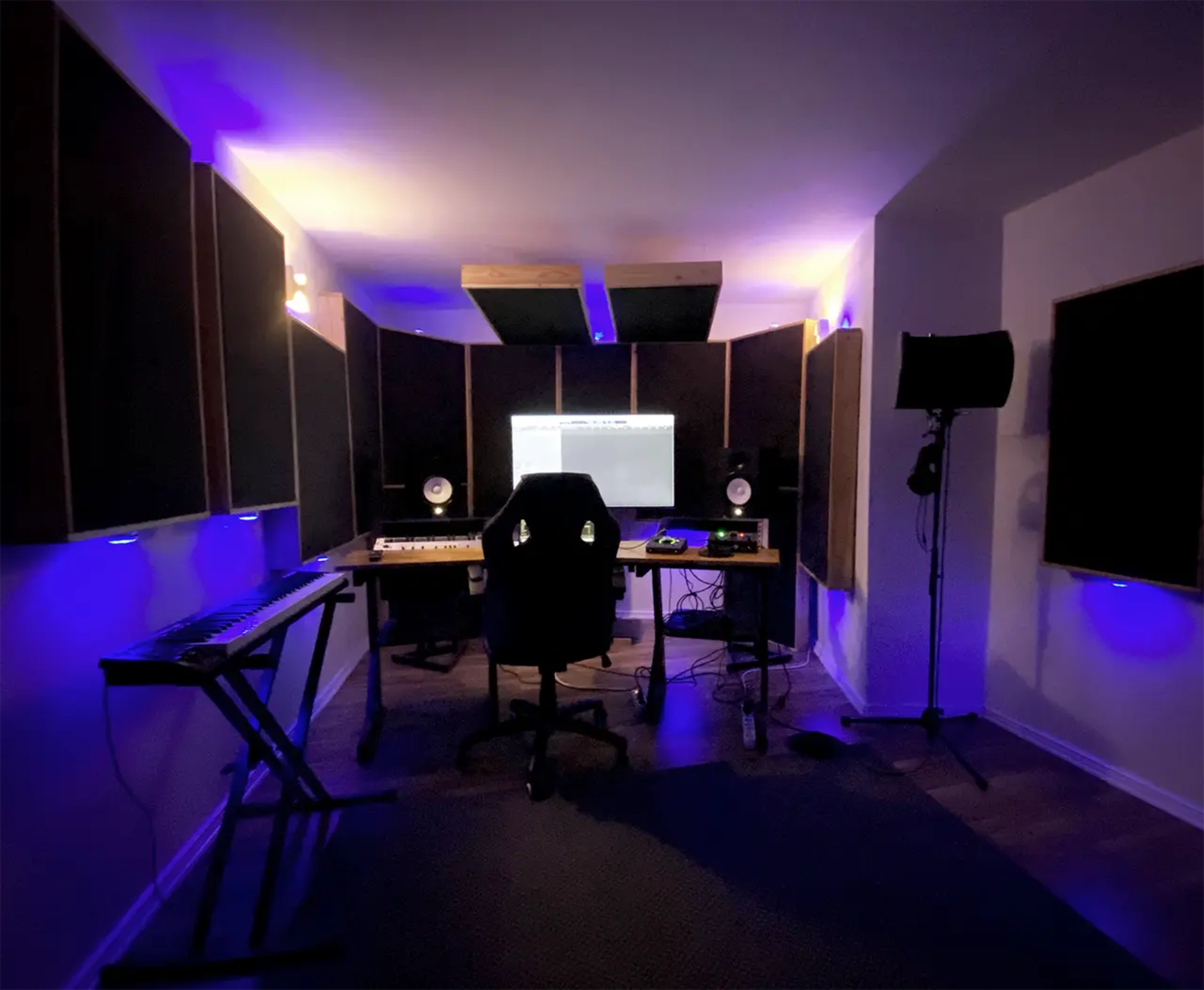 The image shows a music studio with a keyboard, a desk with recording equipment, a chair, and soundproof panels illuminated by purple lighting.