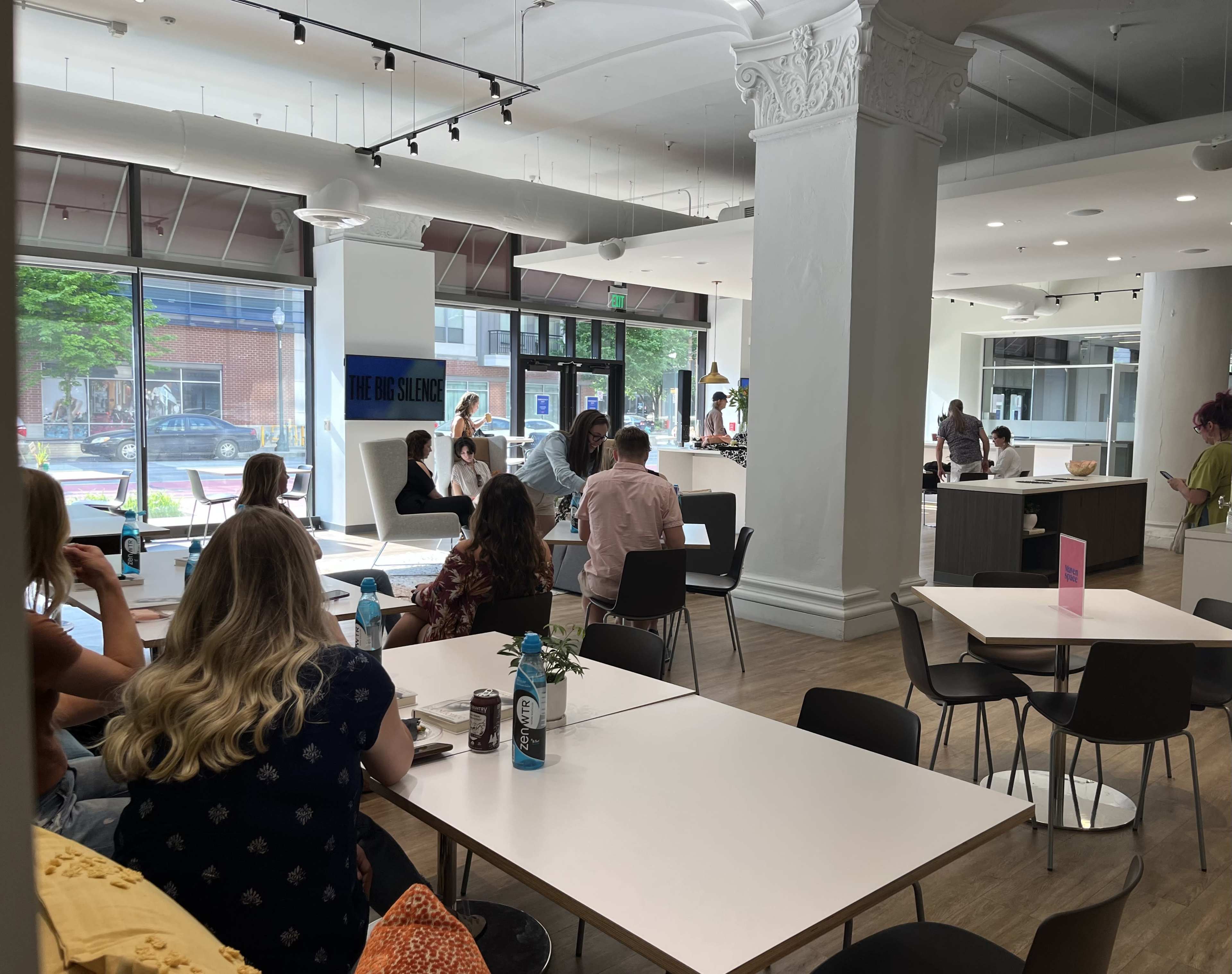 A modern café with a mix of customers seated at tables and a staff member preparing food in a bright, open space with large windows.