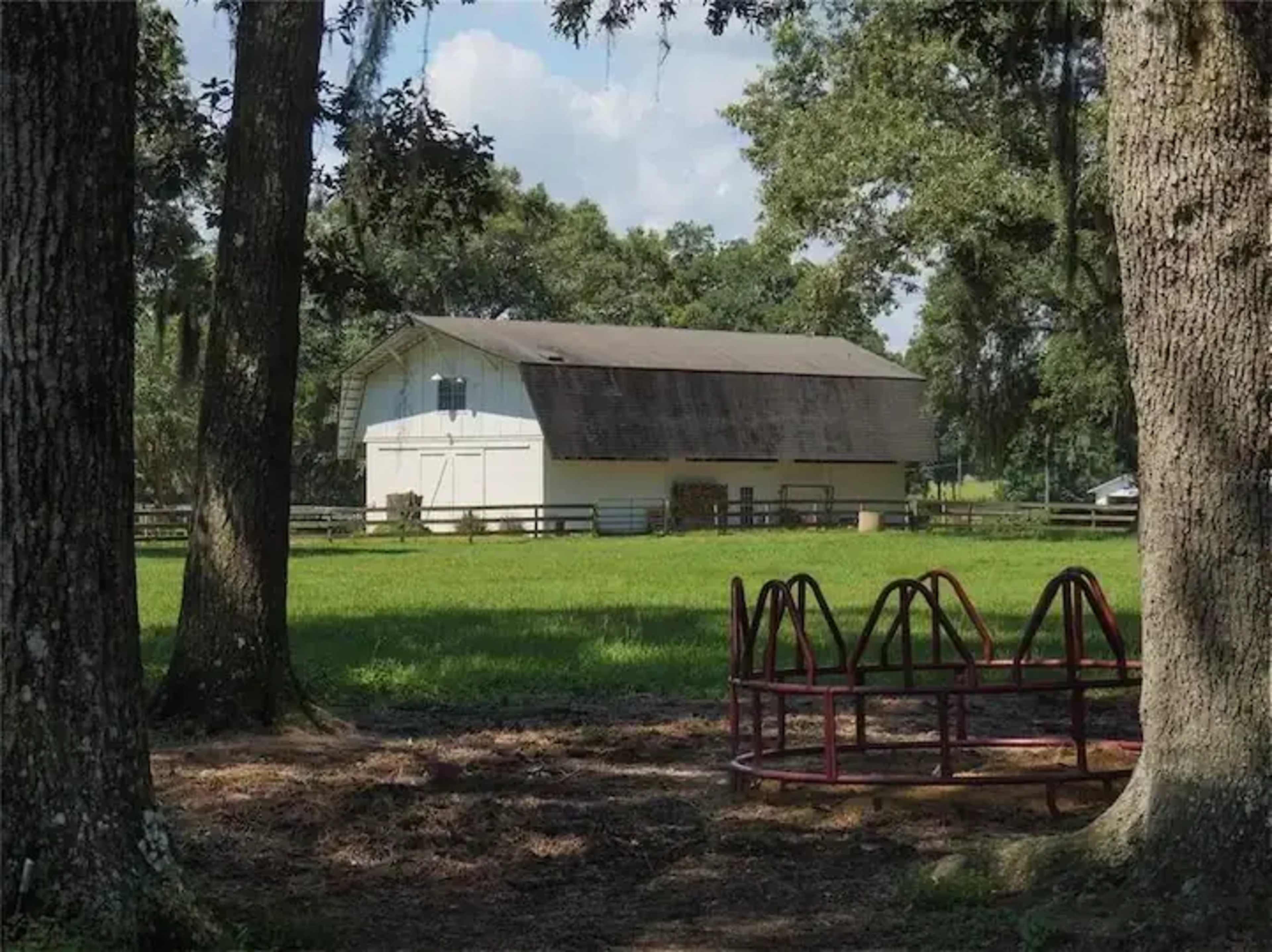 A white barn stands in a grassy field, surrounded by trees, with a playground structure in the foreground.