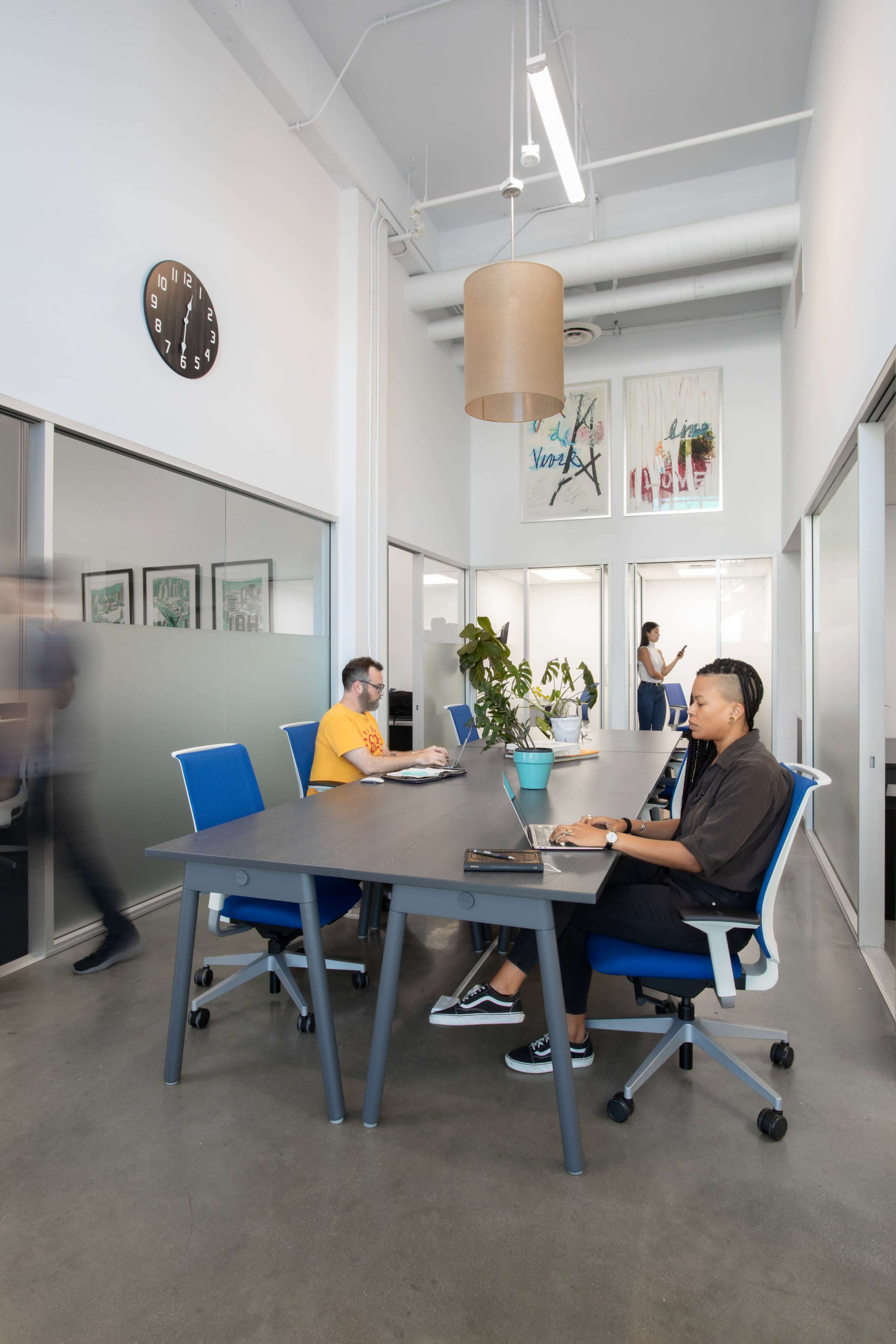 Two individuals work at a large conference table in a modern office space, with a third person visible in the background and a clock on the wall.