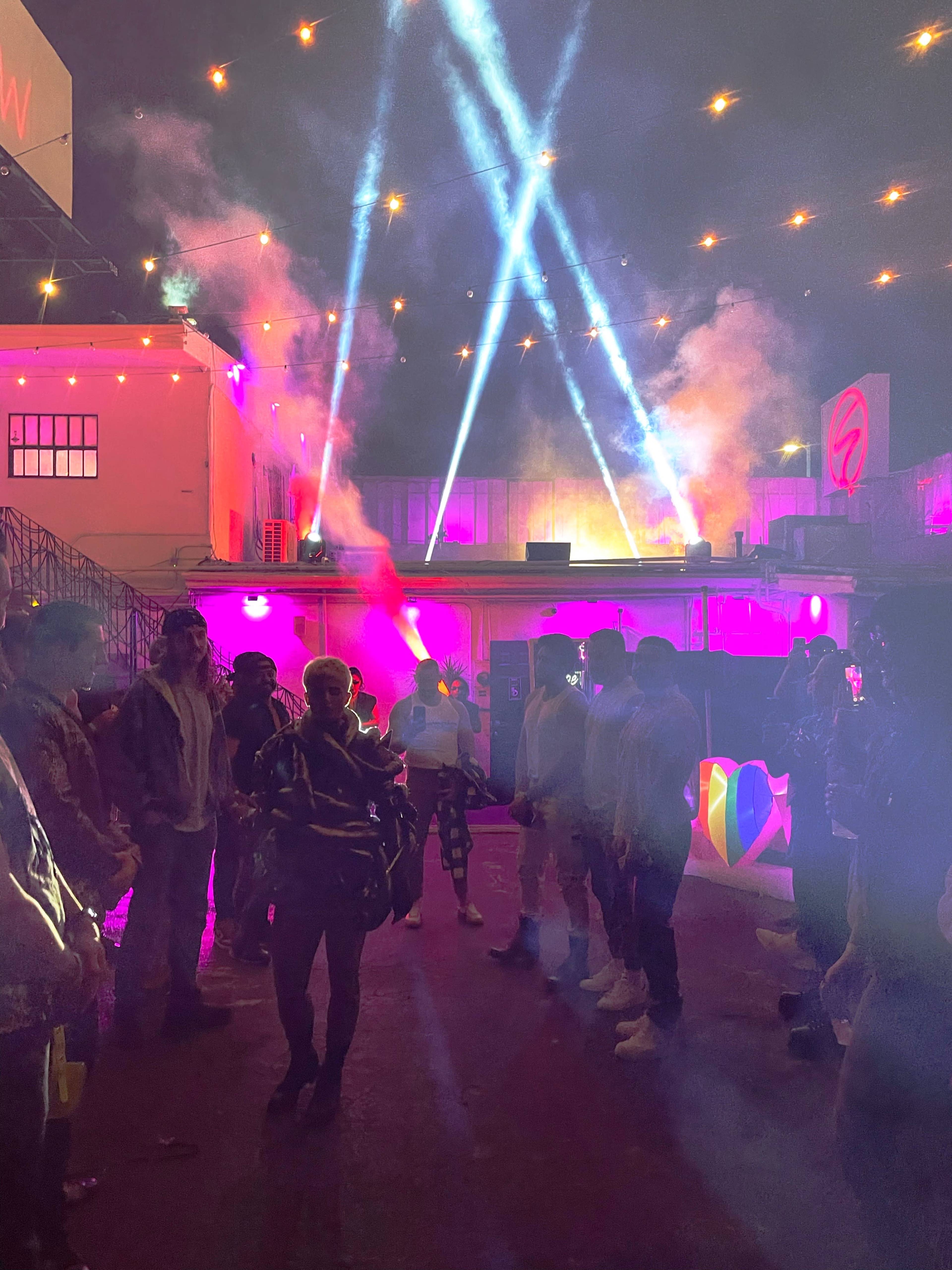 A crowd of people stands in a lit outdoor space with colorful lights and fog effects, while beams of light shine down from above.