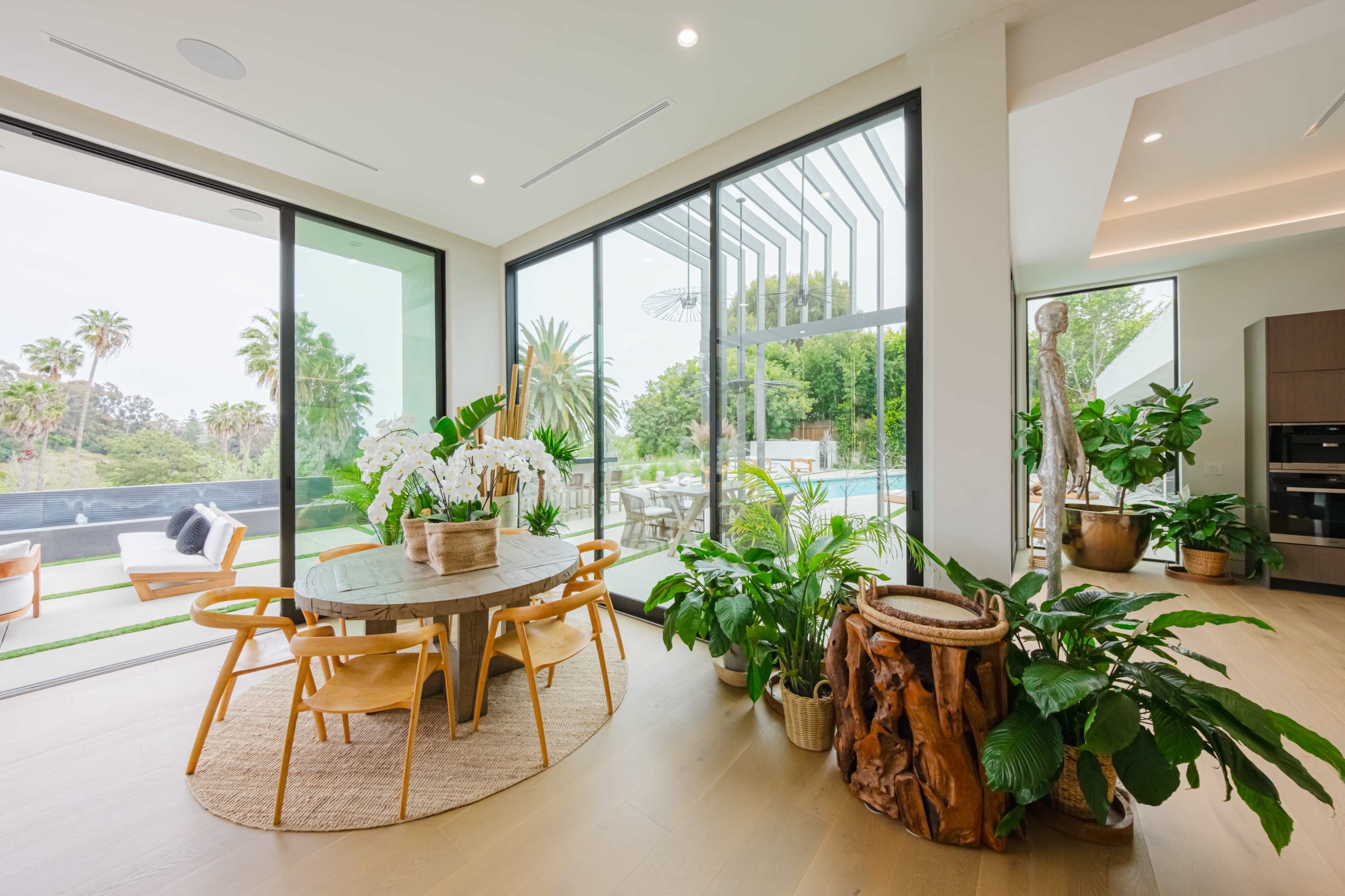 A bright, modern dining area features a round wooden table surrounded by stylish chairs, with large windows providing a view of a landscaped outdoor space and pool.