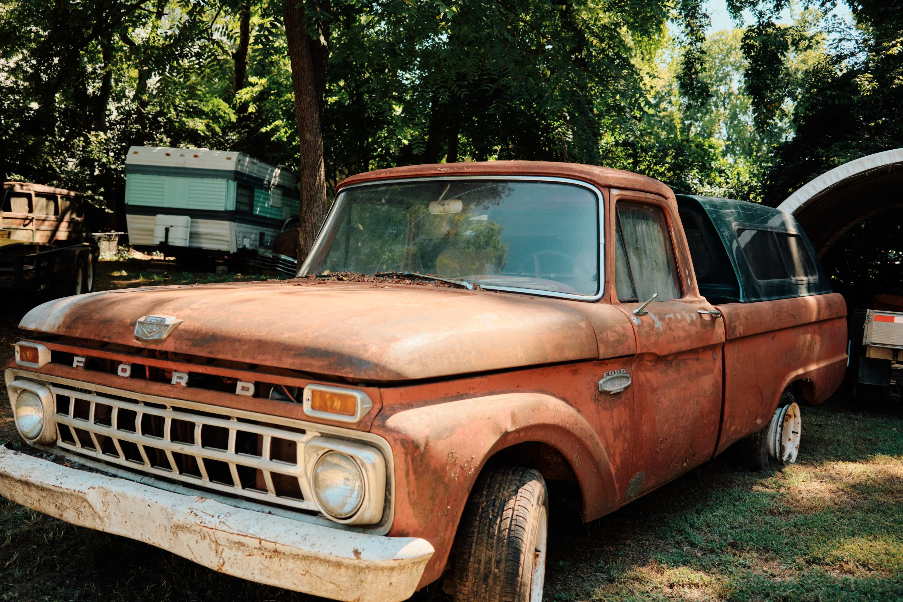 An old, rusty Ford pickup truck is parked on grass under a canopy of trees.