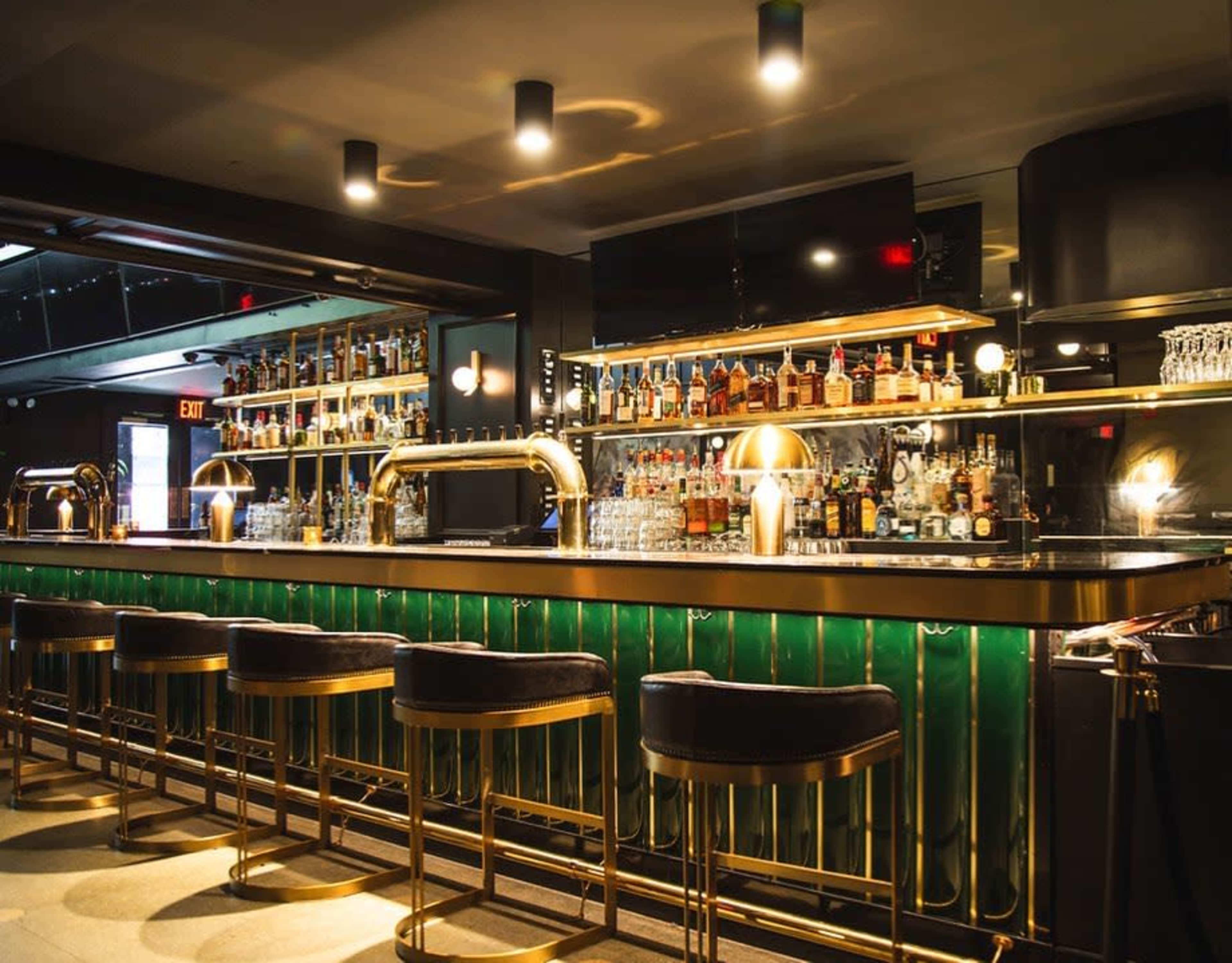 The image shows a modern bar with a polished countertop, backed by shelves filled with various bottles, and sleek bar stools lined up in front.