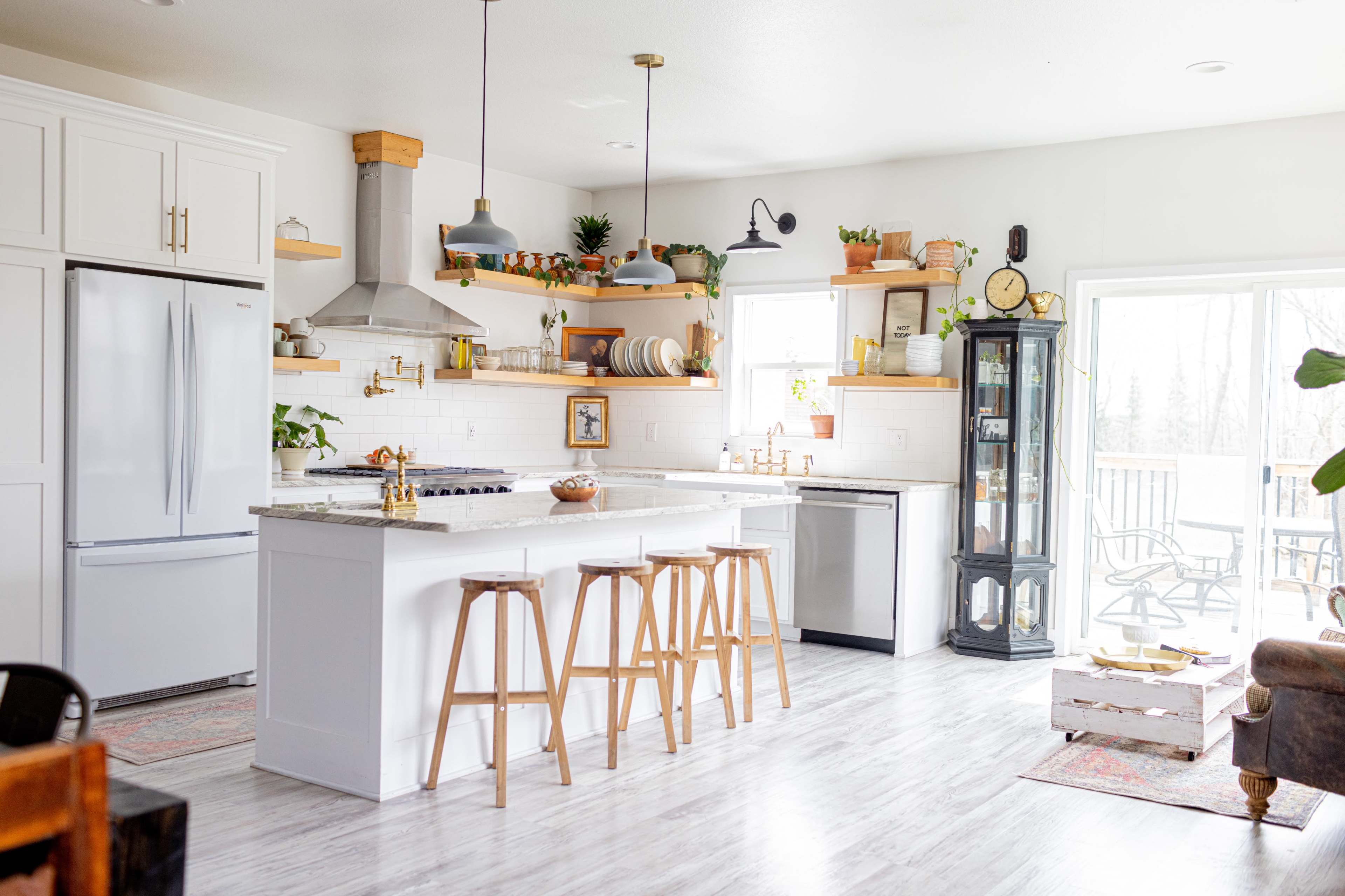 The image shows a modern kitchen with white cabinetry, a large island with wooden stools, and a mix of decorative plants and utensils on the shelves.