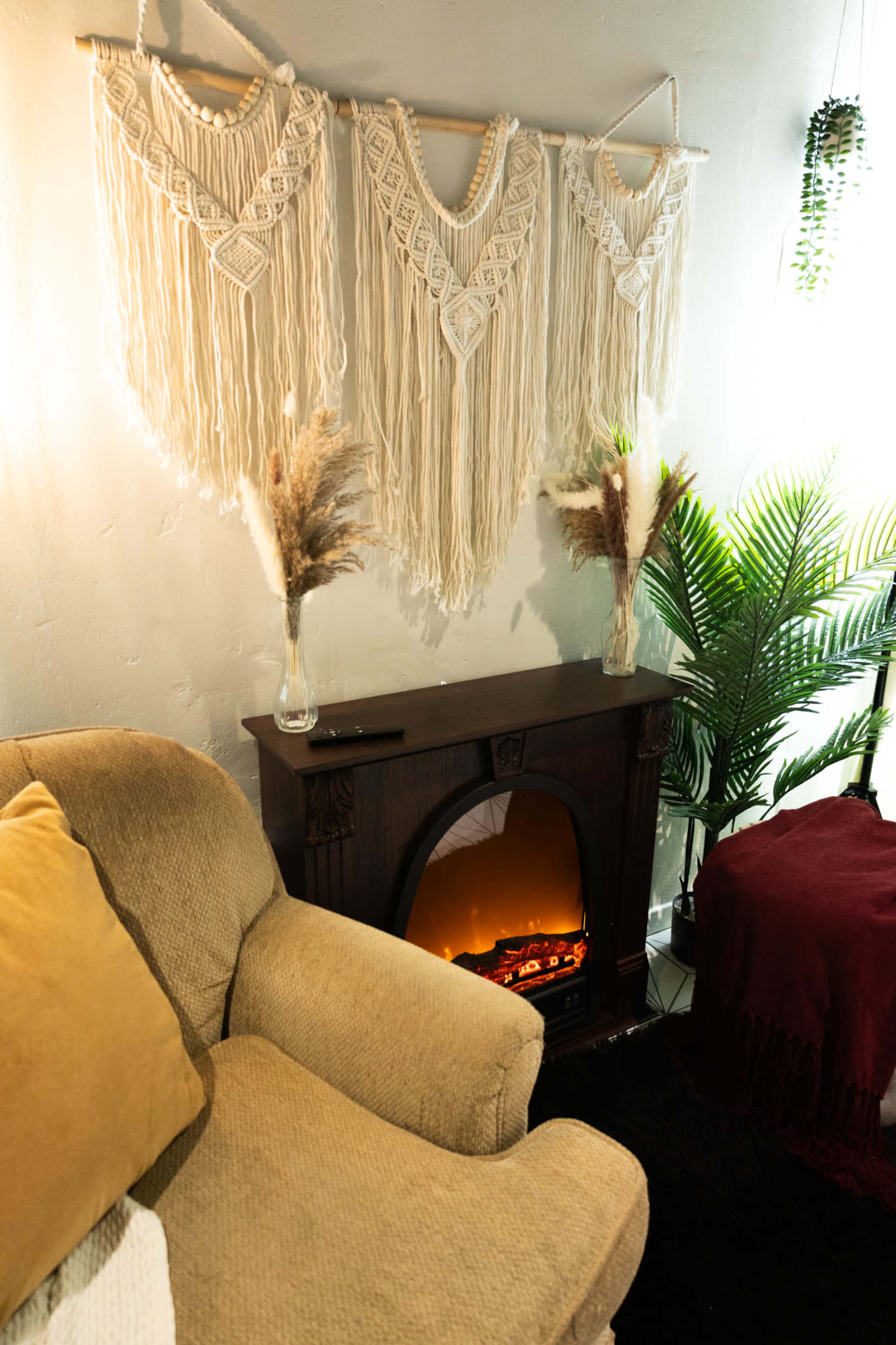 The image features a cozy living space with a beige couch, a decorative macramé wall hanging, and a faux fireplace, complemented by potted plants and decorative pampas grass.