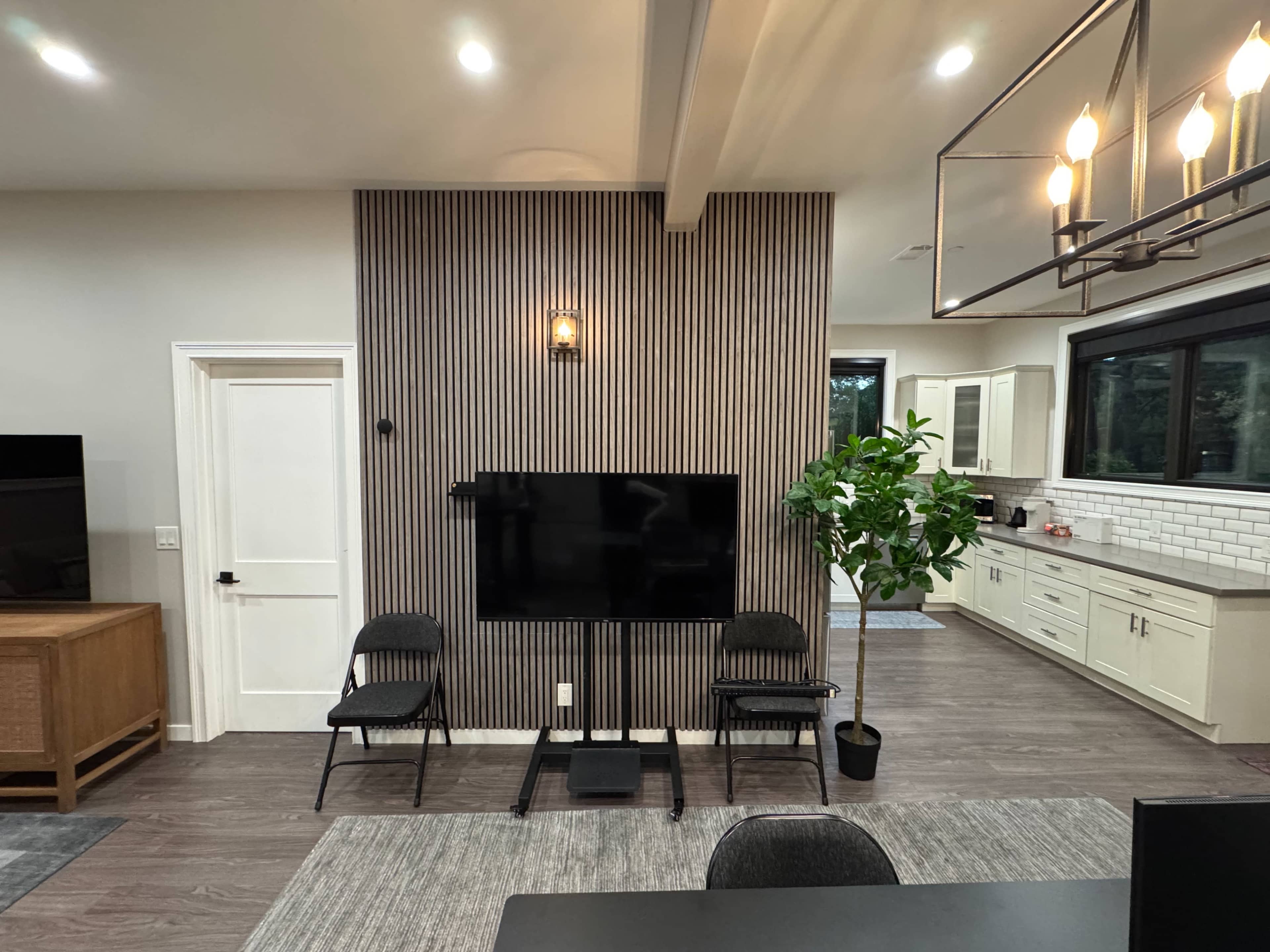 The image shows a modern living area with a TV mounted on a slatted feature wall, two black chairs, a potted plant, and a kitchen visible in the background.