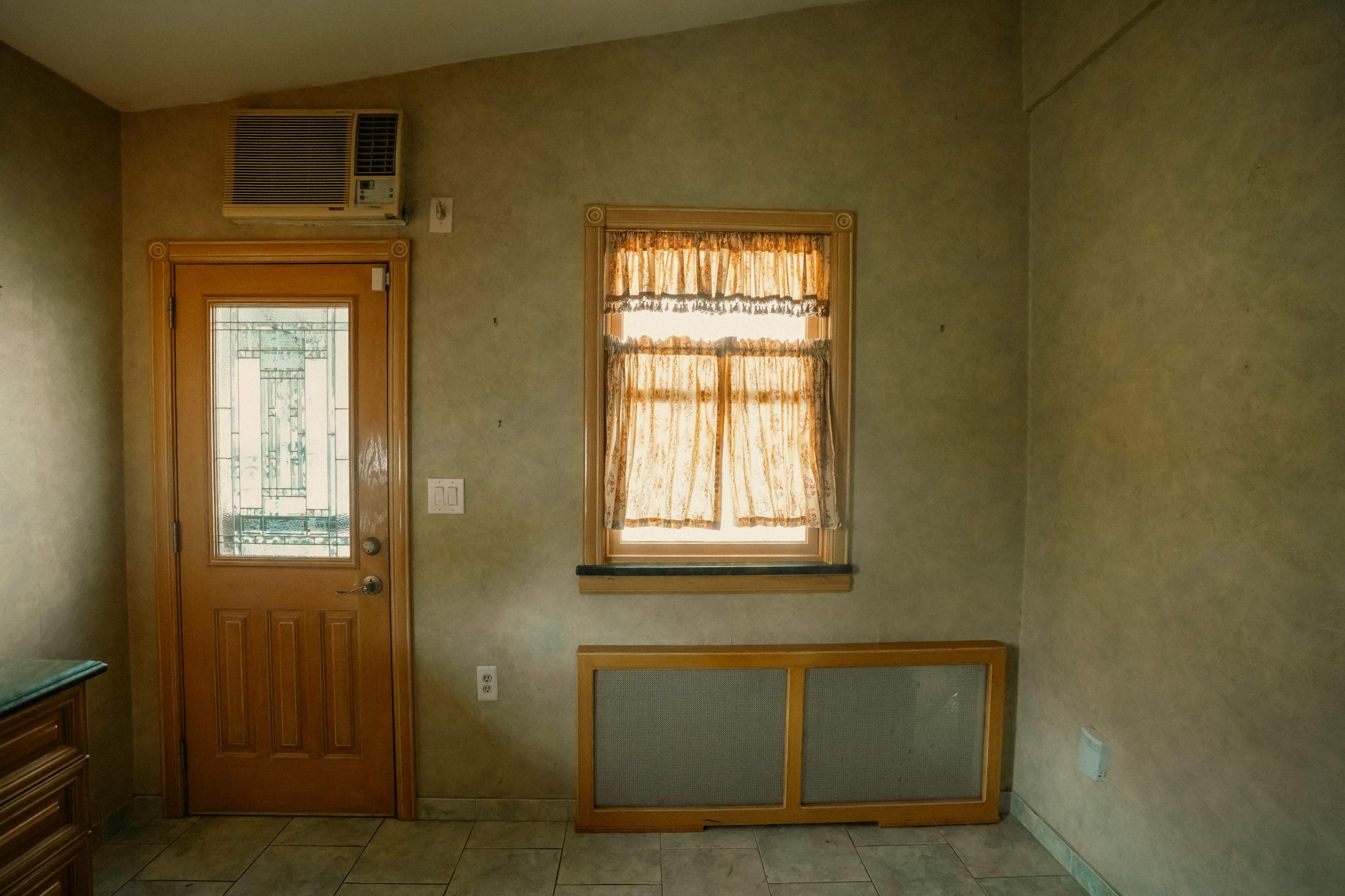 The image shows a small, empty room with a wooden door, a window with sheer curtains, and an air conditioning unit mounted on the wall.